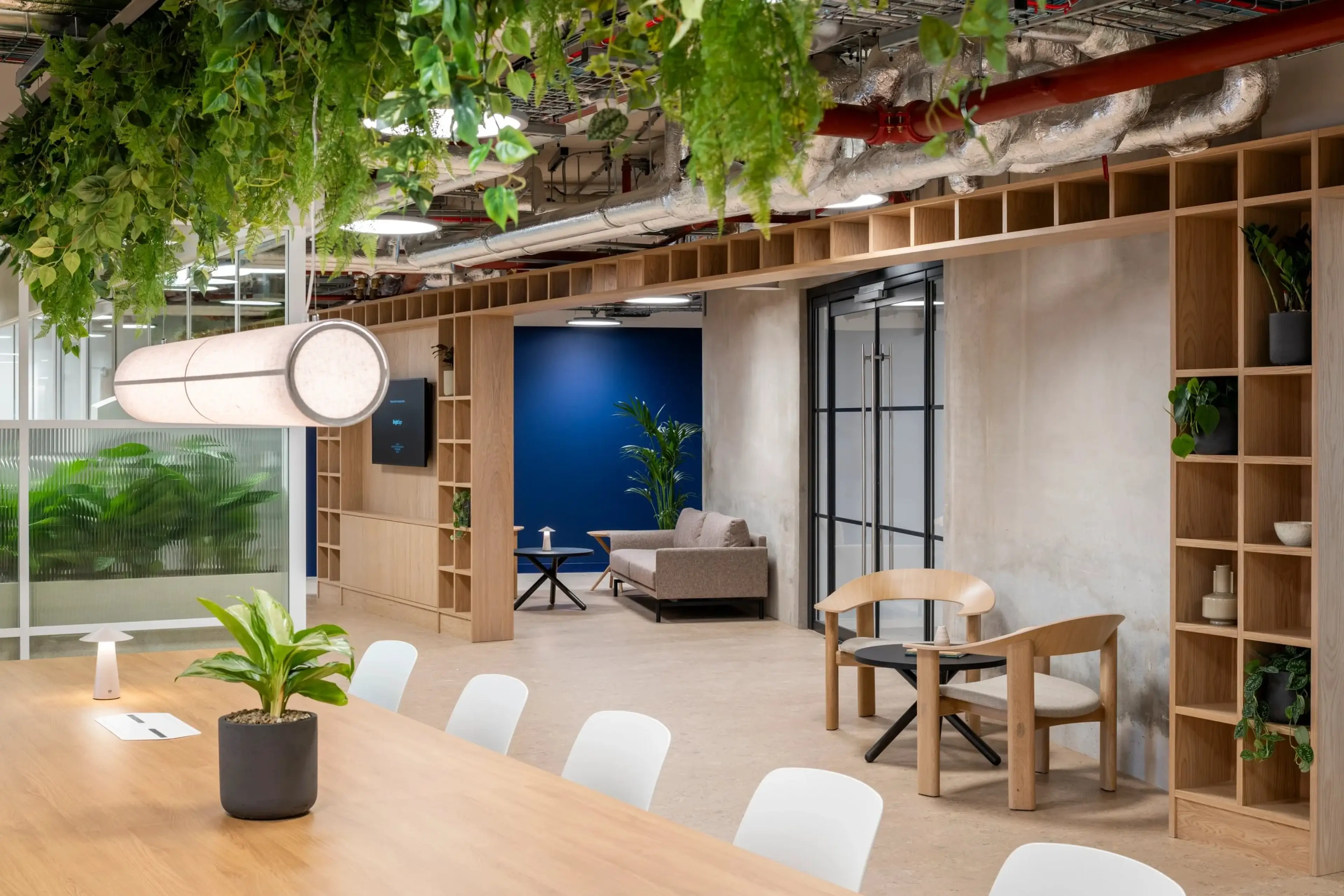 Modern office space with wooden furniture, hanging plants, and a large table. A cozy lounge area with a sofa and chairs is visible in the background.
