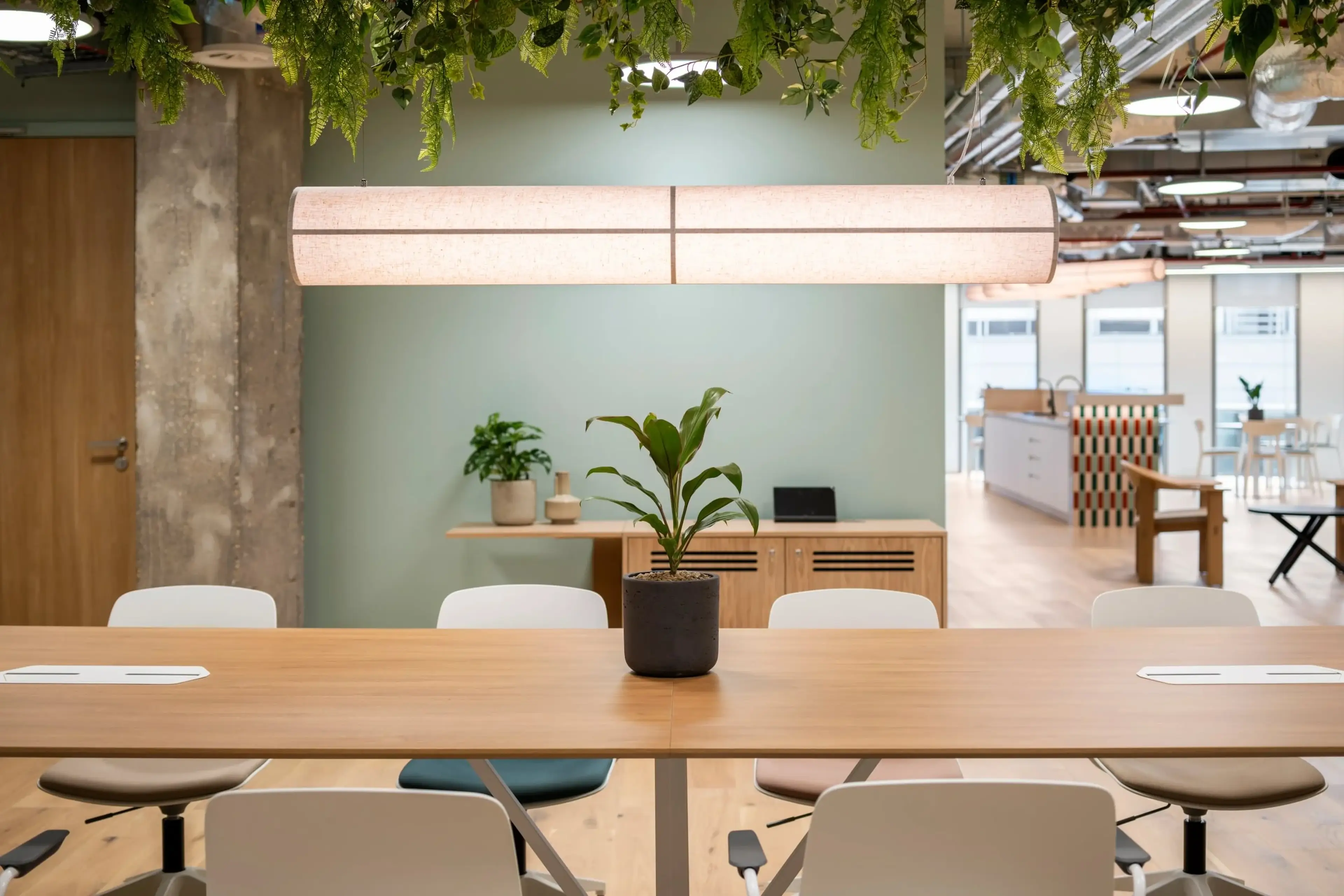 Modern office with wooden table, white chairs, hanging plants, and ambient lighting. Potted plant centerpiece on table. Open, airy design.