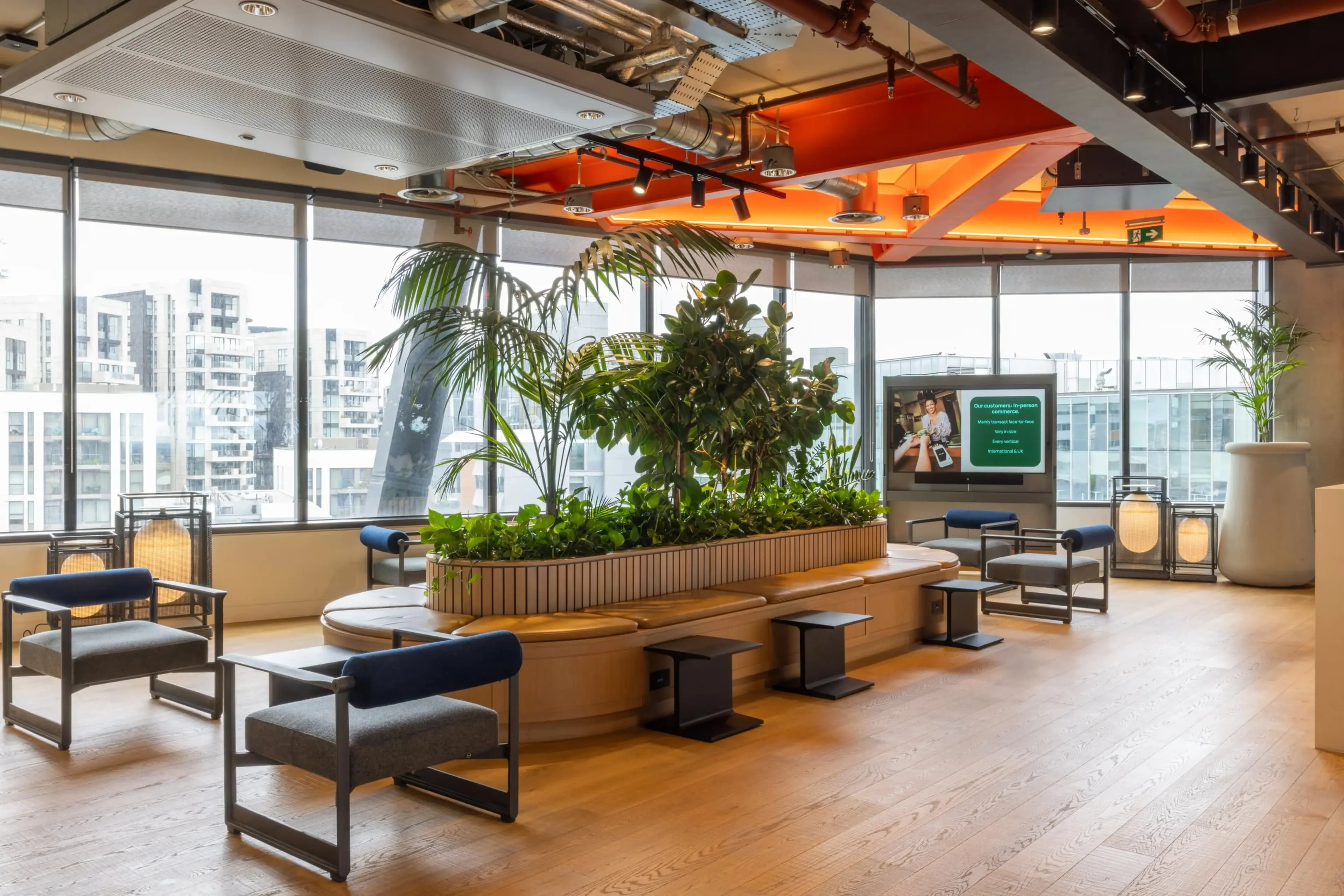 Modern office lounge with large windows, greenery, wooden floor, stylish chairs, and a digital screen. Bright, airy, and inviting space.
