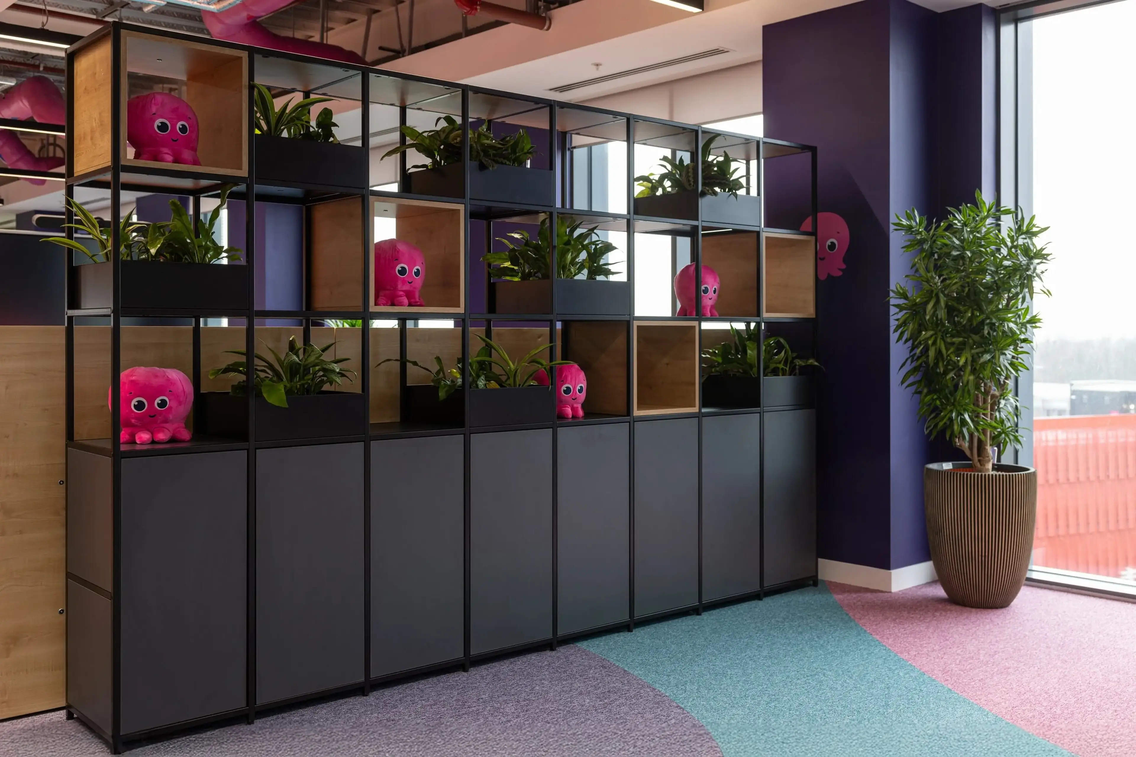 Office space with colorful flooring, plant-filled shelves, and pink plush toys. Large windows and a potted plant add brightness and greenery.