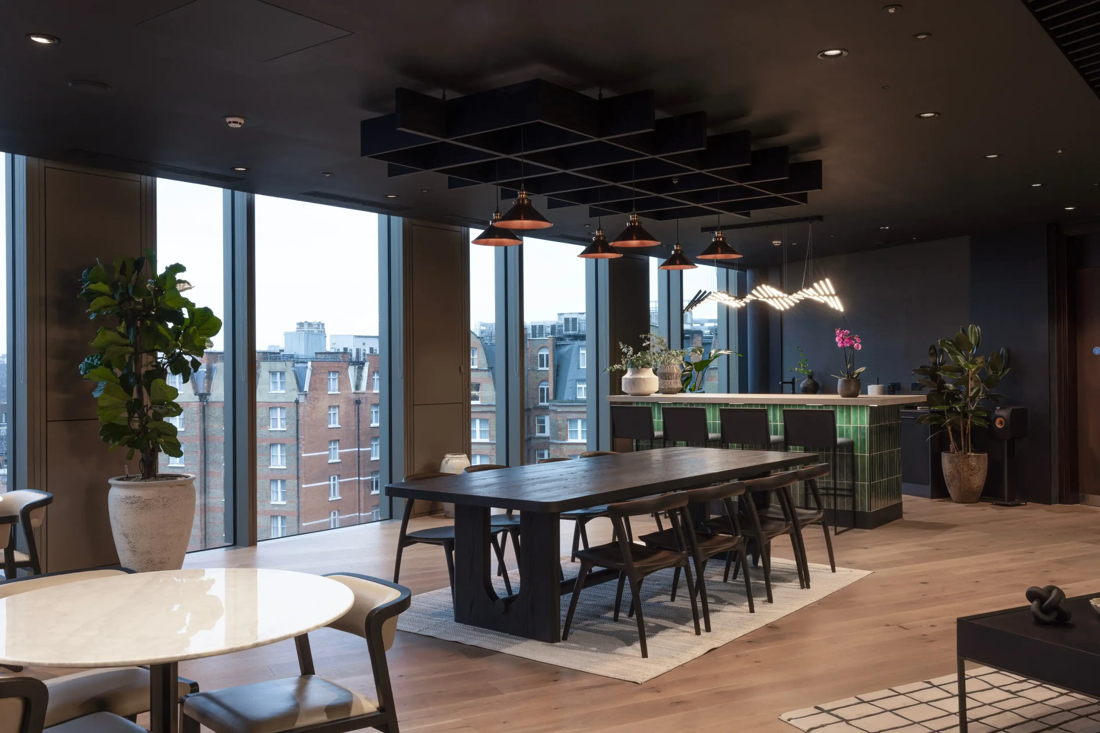 A sophisticated office social space with a long dark wood dining table, surrounded by black chairs. The background features a bar area with green tiled cladding, pendant lighting, and large windows offering city views.