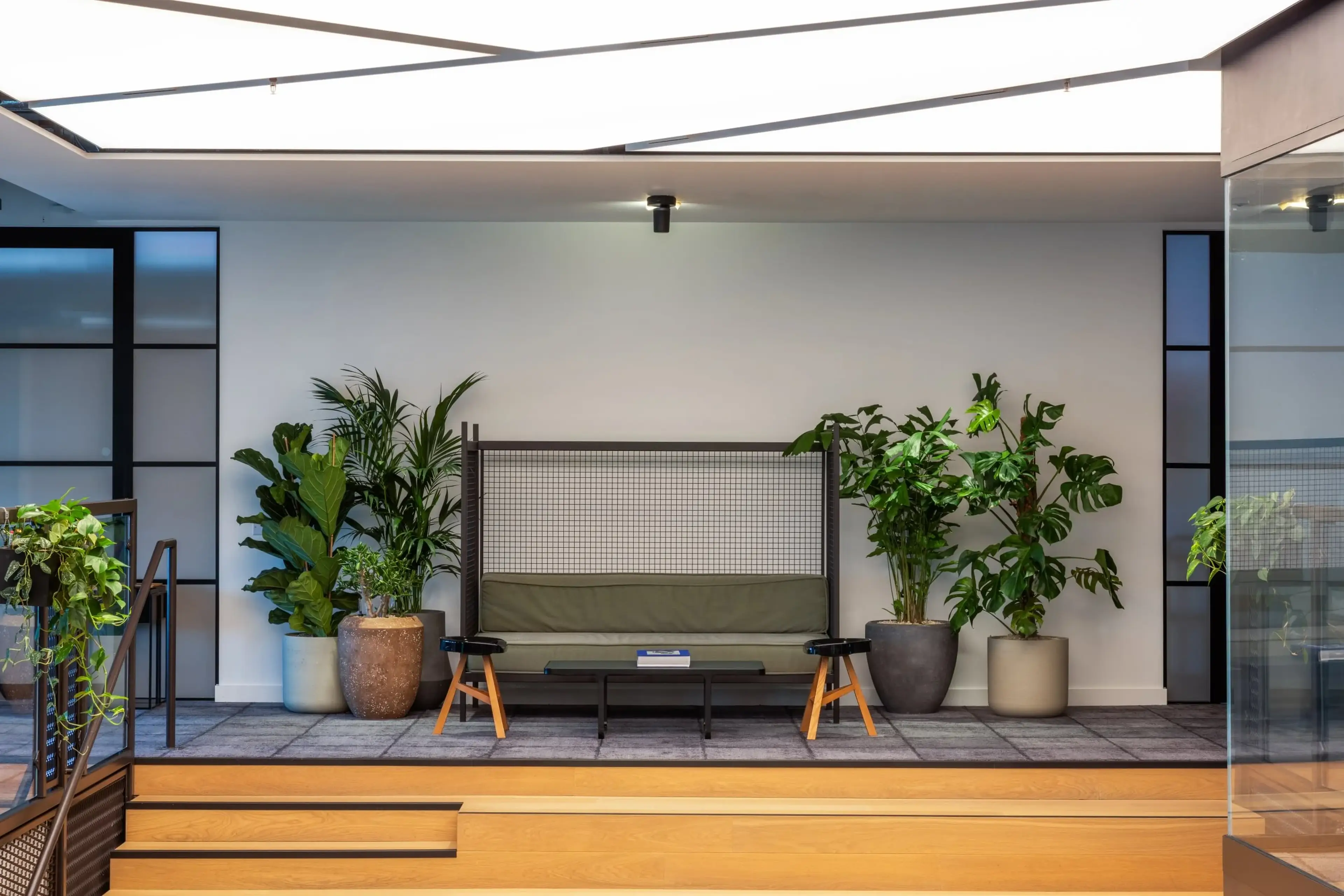Modern indoor seating area with a green cushioned bench, wooden stools, large potted plants, and a skylight above.