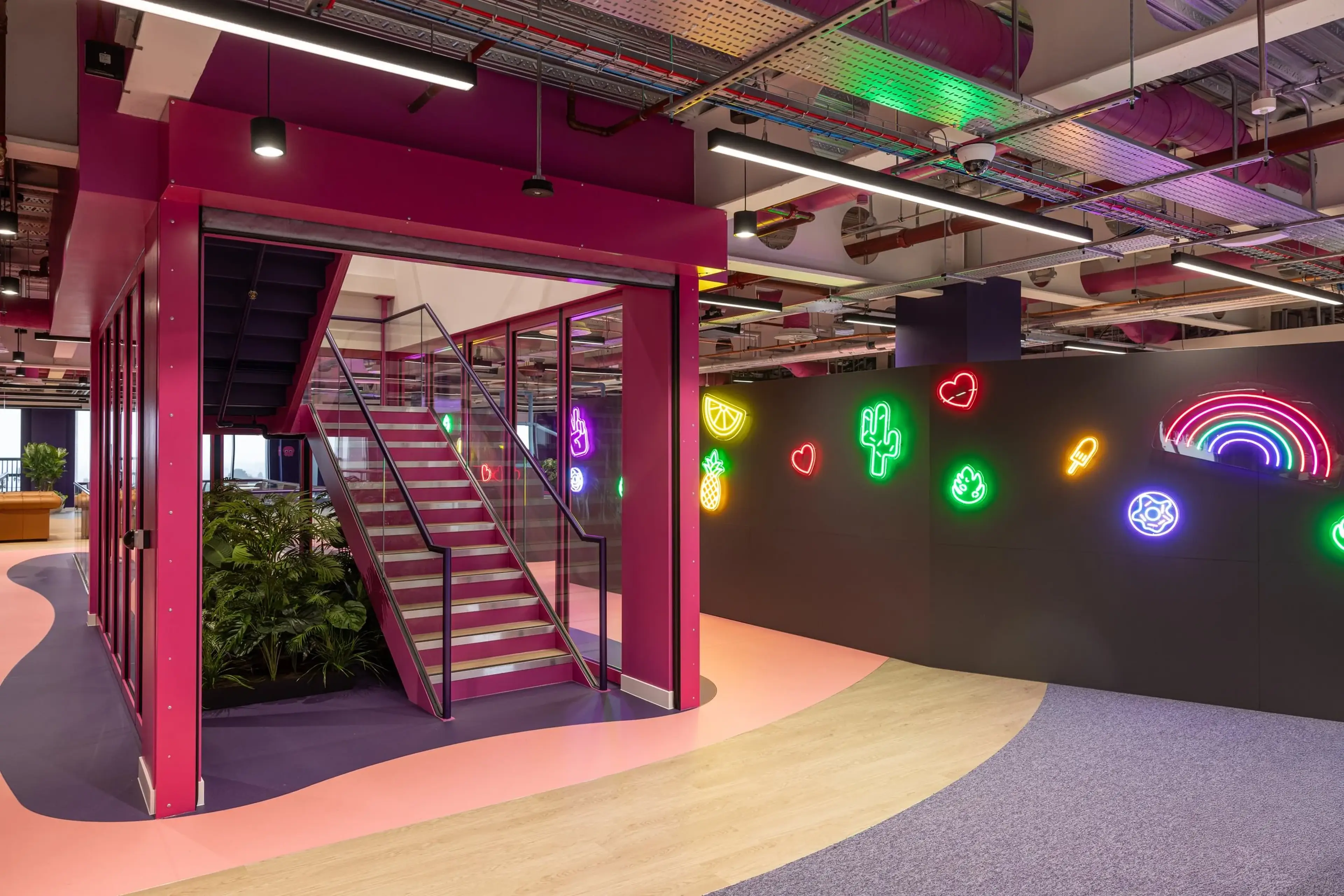 Modern office interior with vibrant neon signs on a black wall, a staircase with pink accents, and colourful flooring.