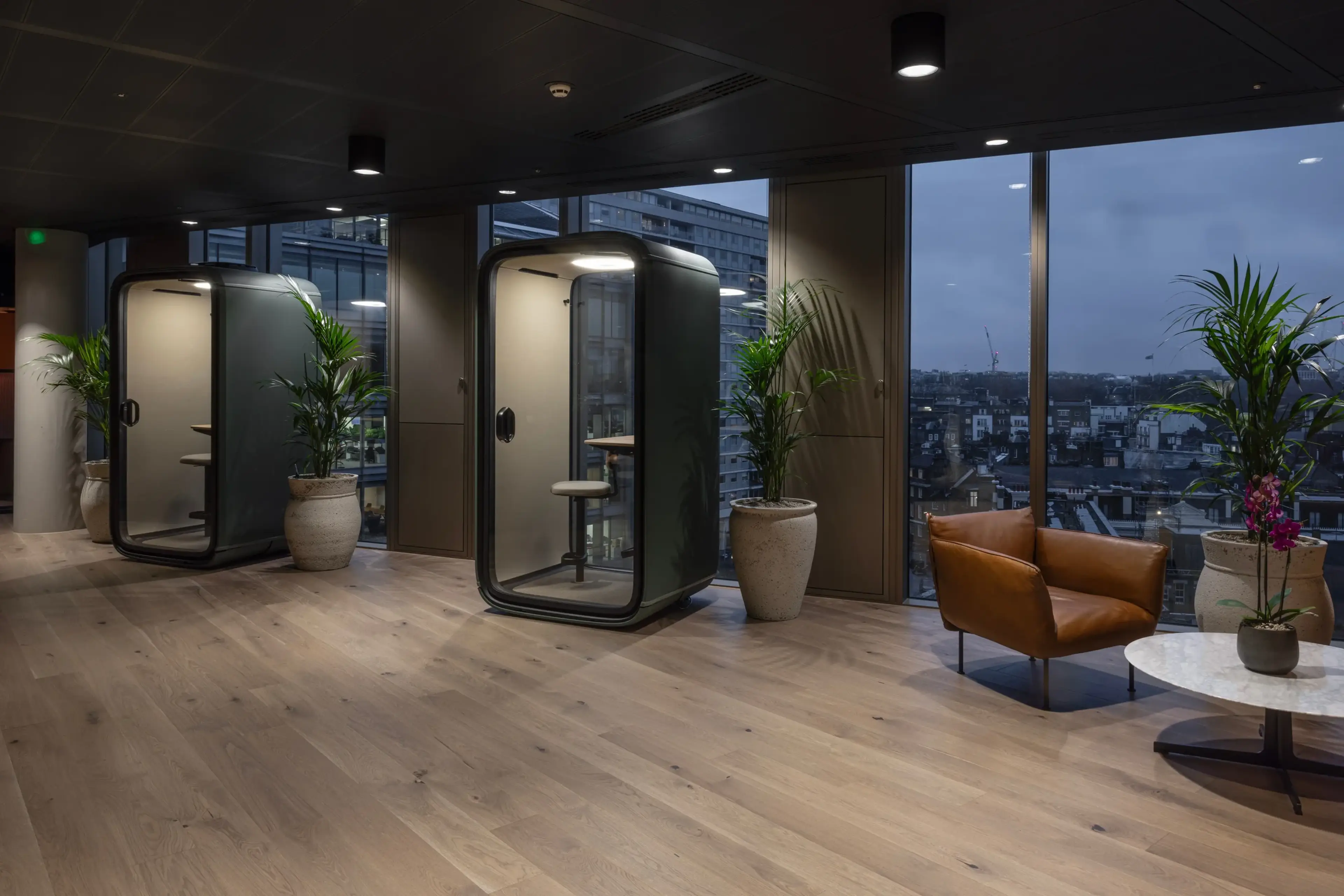 A modern office breakout area featuring two sleek acoustic phone booths with glass doors, set against floor-to-ceiling windows offering a cityscape view. The space is complemented by large potted palms in textured stone planters, a tan leather lounge chair, and a round marble coffee table with an orchid.