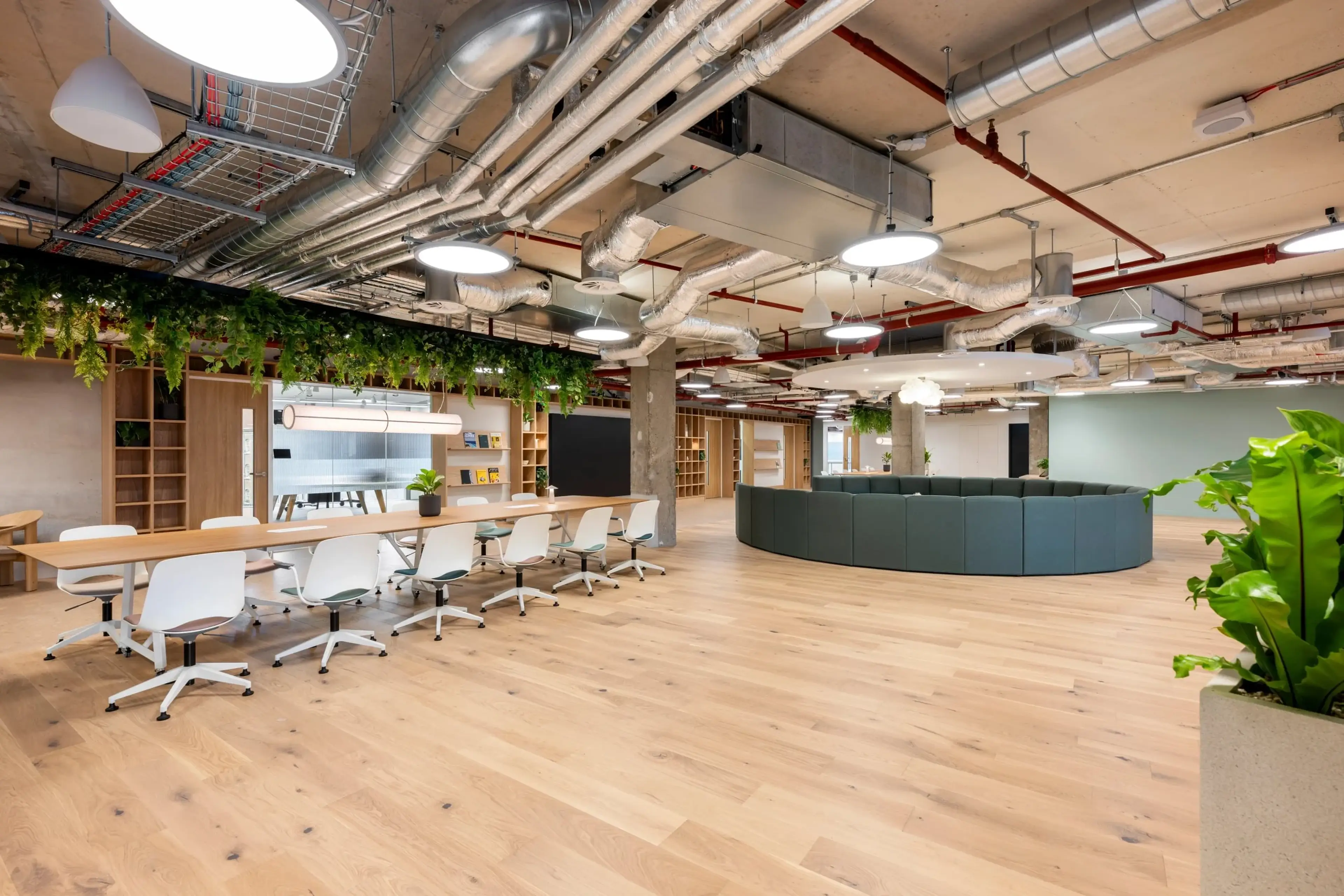 Modern office space with wood flooring, a long table with chairs, industrial ceiling, plants, and a circular seating area.
