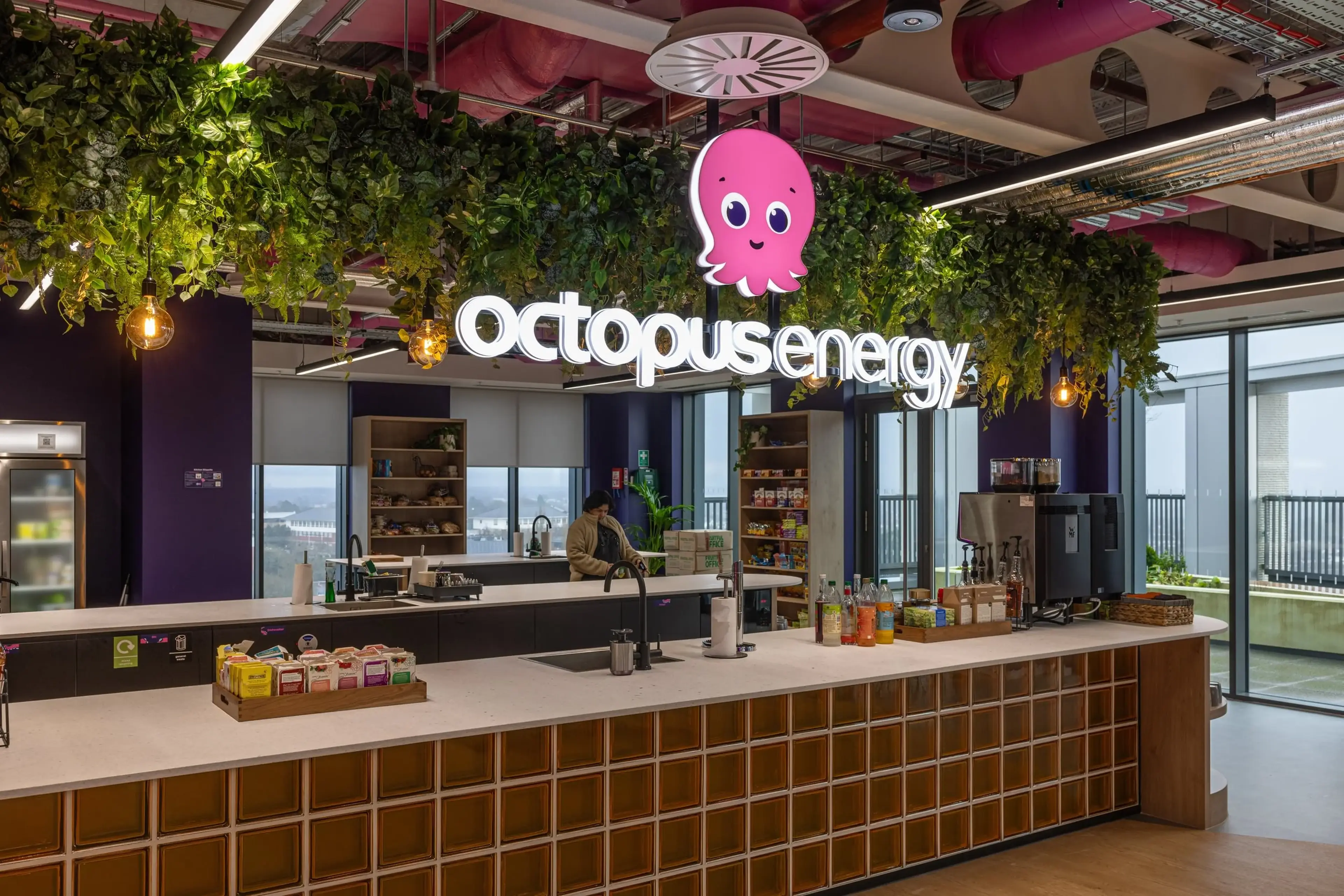 Modern office kitchen with "Octopus Energy" sign, hanging plants, snacks, drinks, and large windows offering a city view.