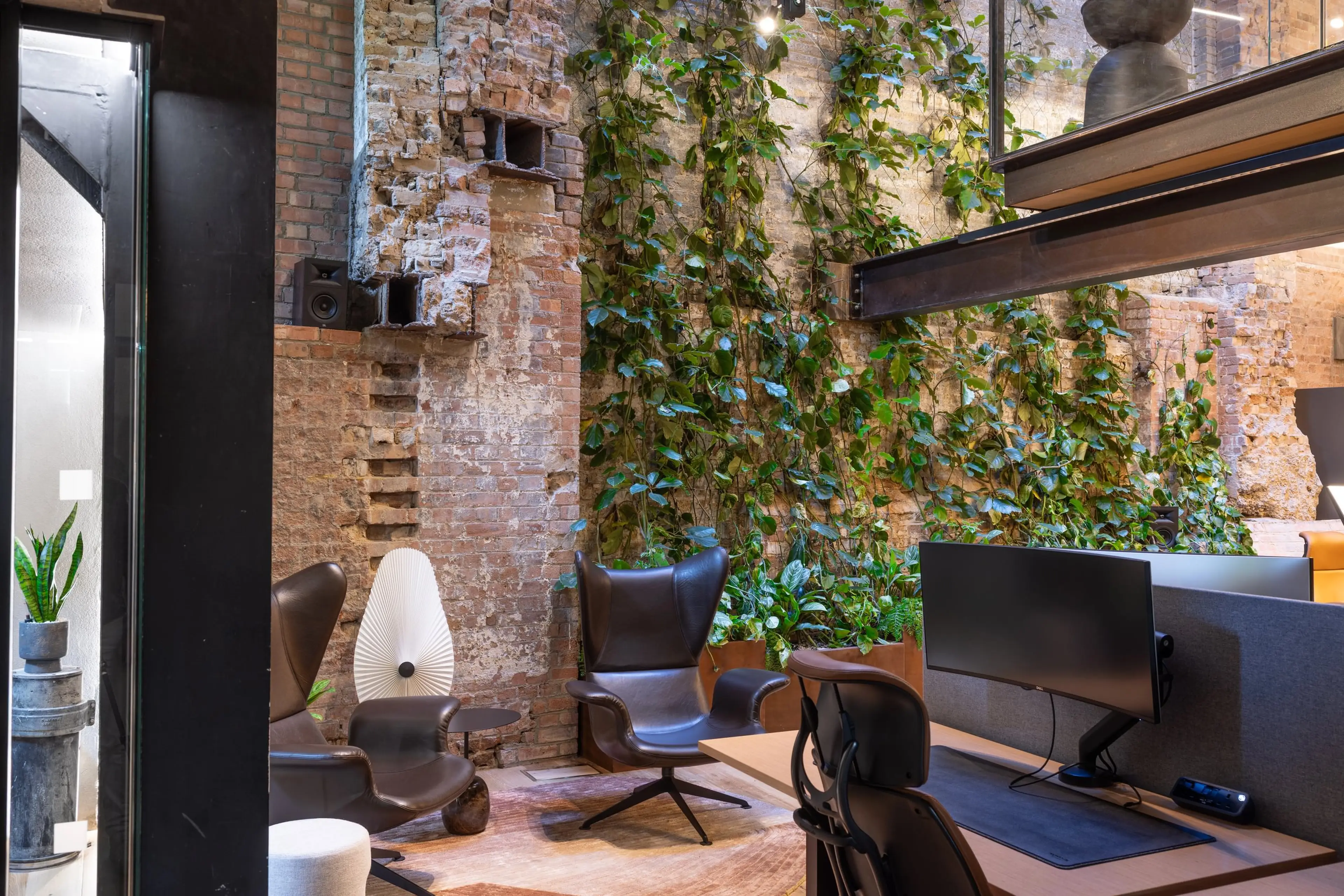 Modern office with exposed brick walls, vertical garden, sleek chairs, and a desk with a computer monitor. Natural and industrial design elements.