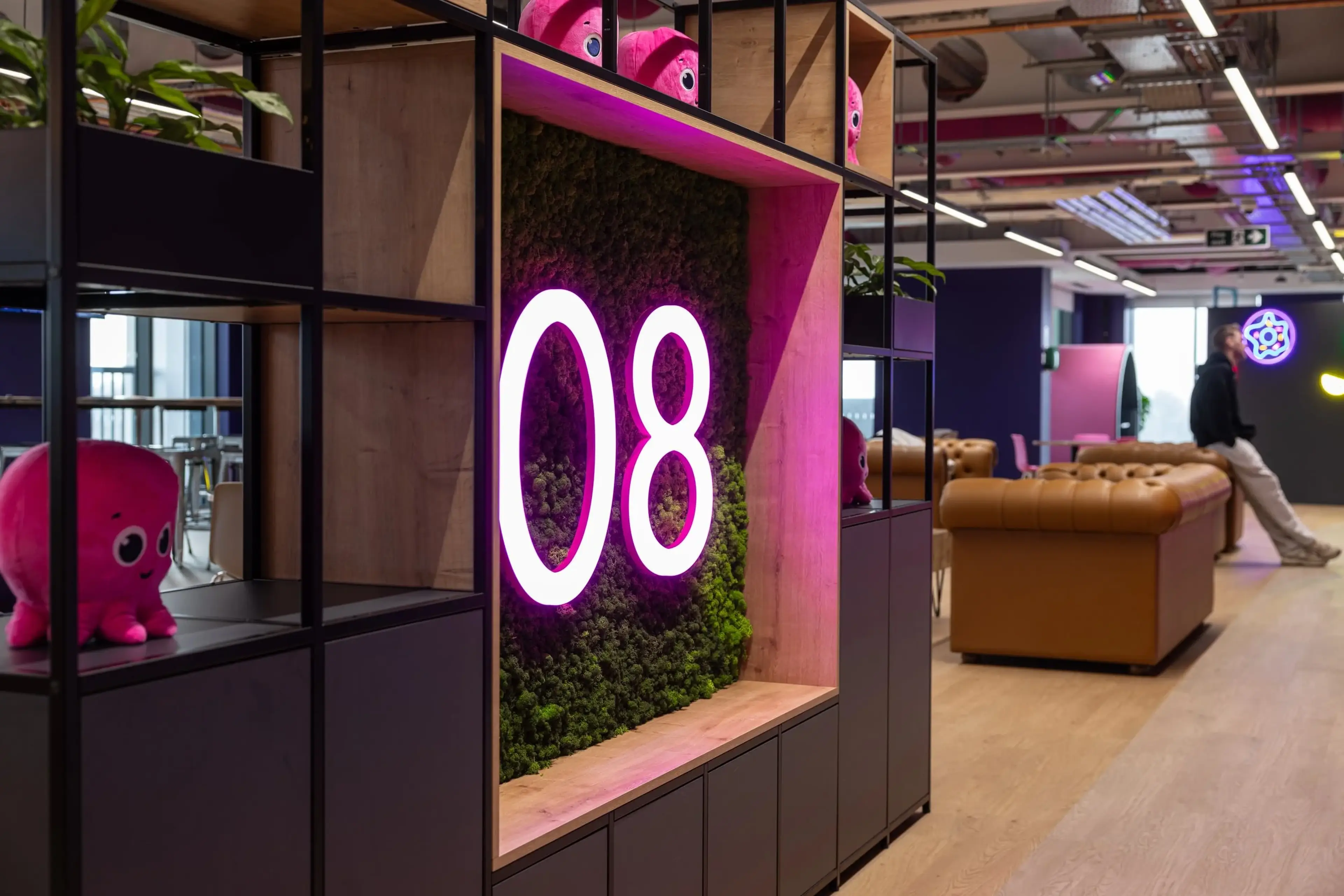 Modern office space with a neon "08" sign on a moss wall, pink plush toys, brown leather sofas, and open ceiling with exposed ducts.