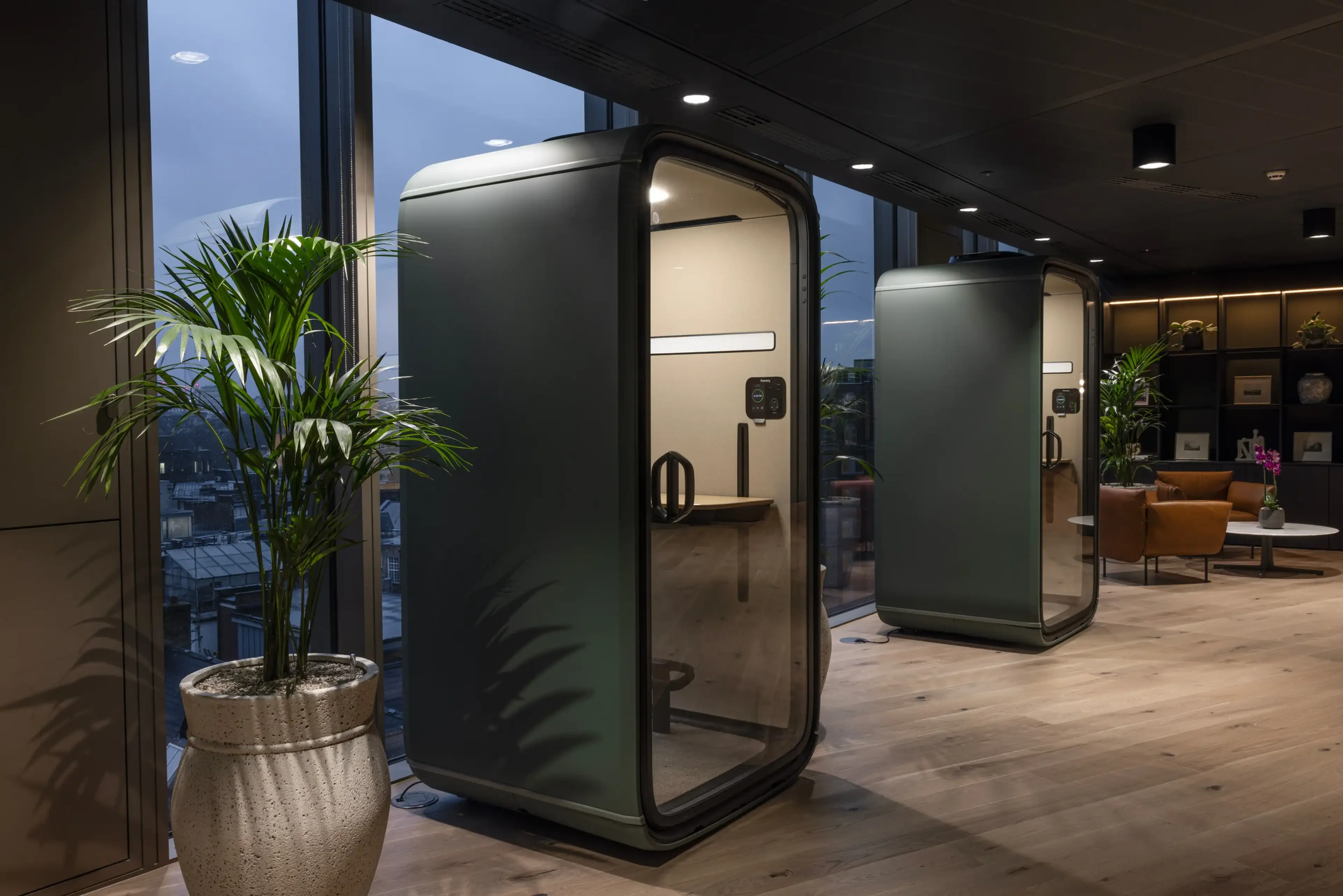 Modern office space with two soundproof pods, large windows, potted office plants, and comfortable seating, creating a sleek and functional environment.