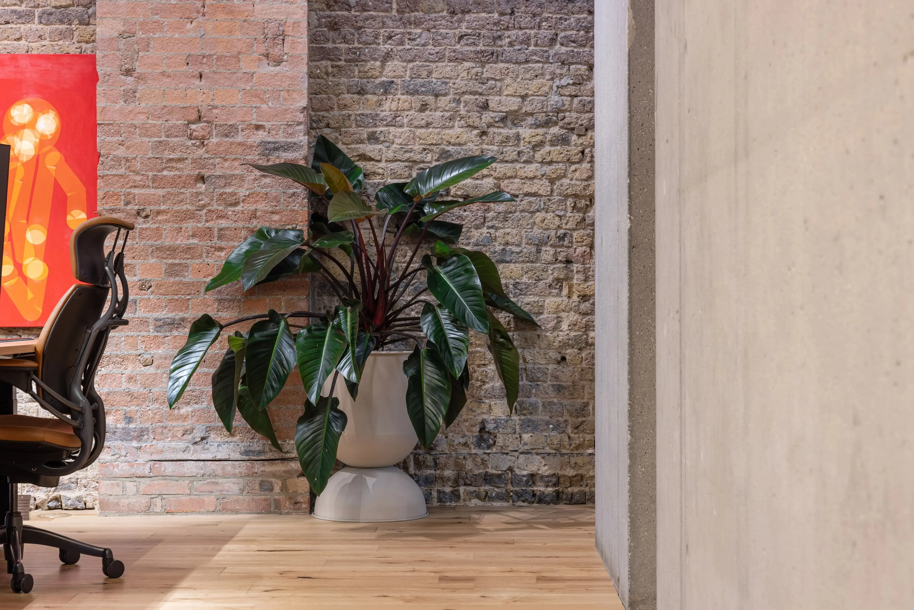 Modern office space with a potted plant, exposed brick wall, wooden floor, and a colorful abstract painting partially visible on the left.