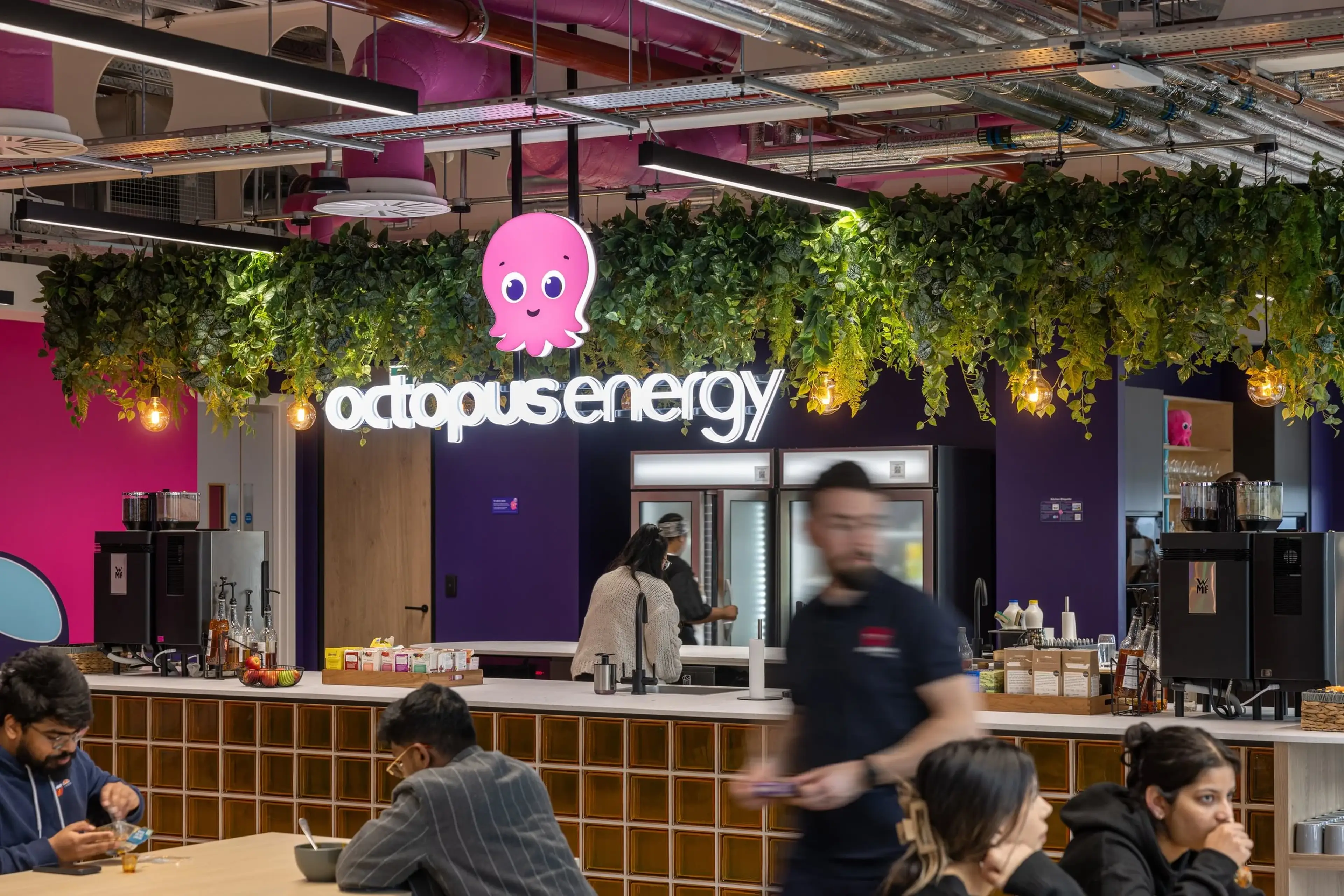 Busy office canteen with people working, featuring a neon "octopus energy" sign and hanging plants above a counter with various items.