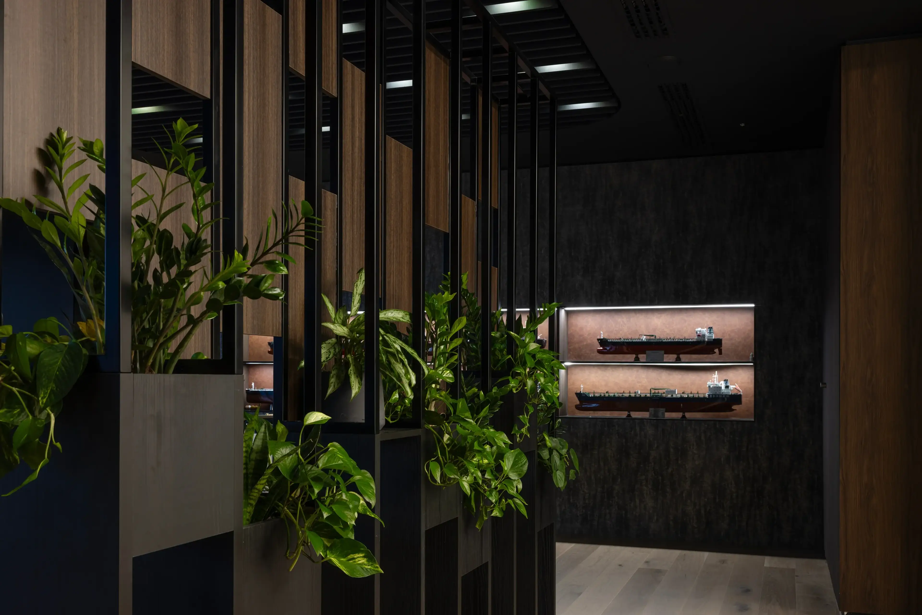 A close-up of a biophilic office divider with vertical black metal frames and wooden panels, featuring integrated planters filled with vibrant green foliage. The background includes illuminated display shelves showcasing ship models.