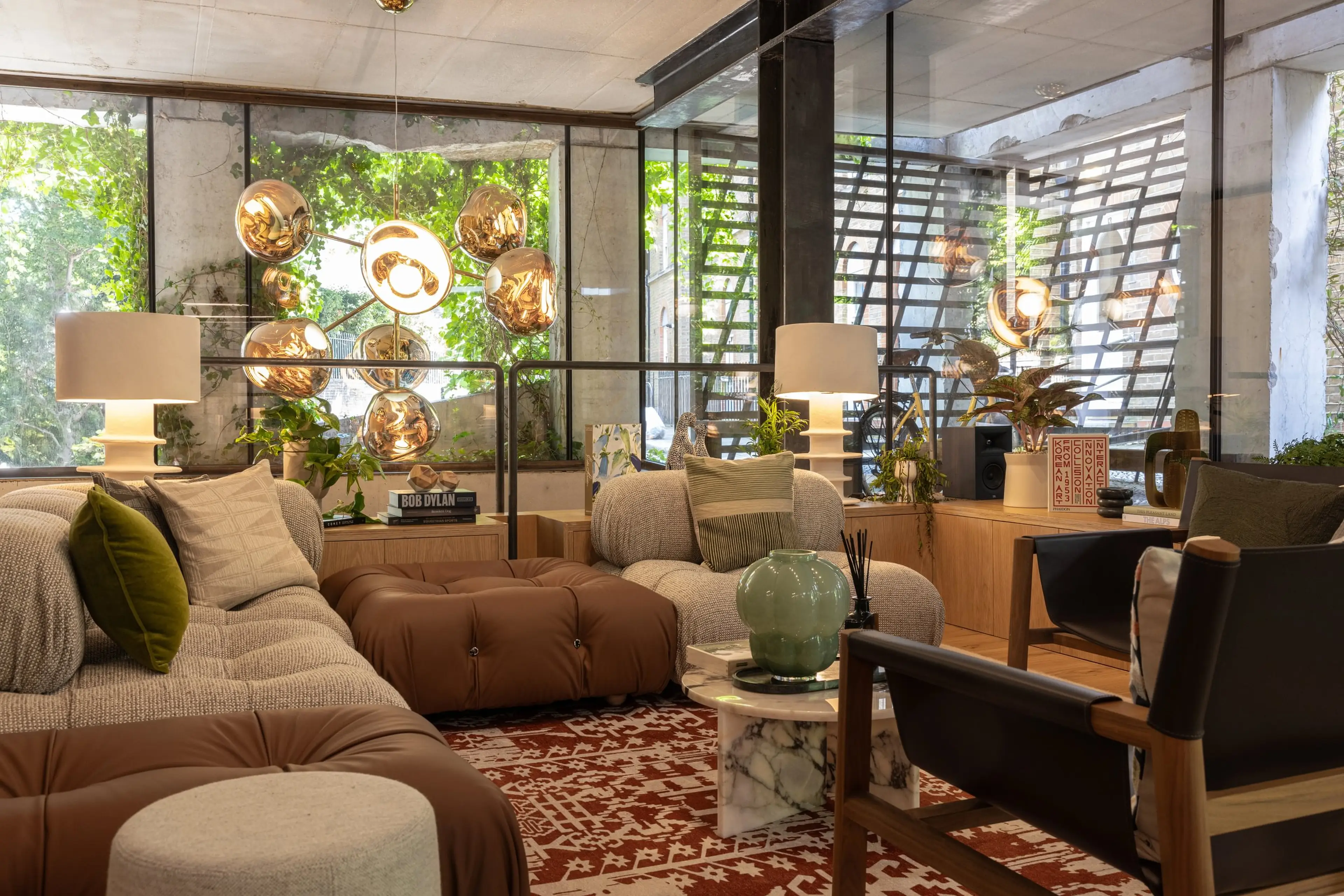 Modern living room with plush seating, a red patterned rug, and spherical pendant lights. Large windows reveal lush greenery outside.