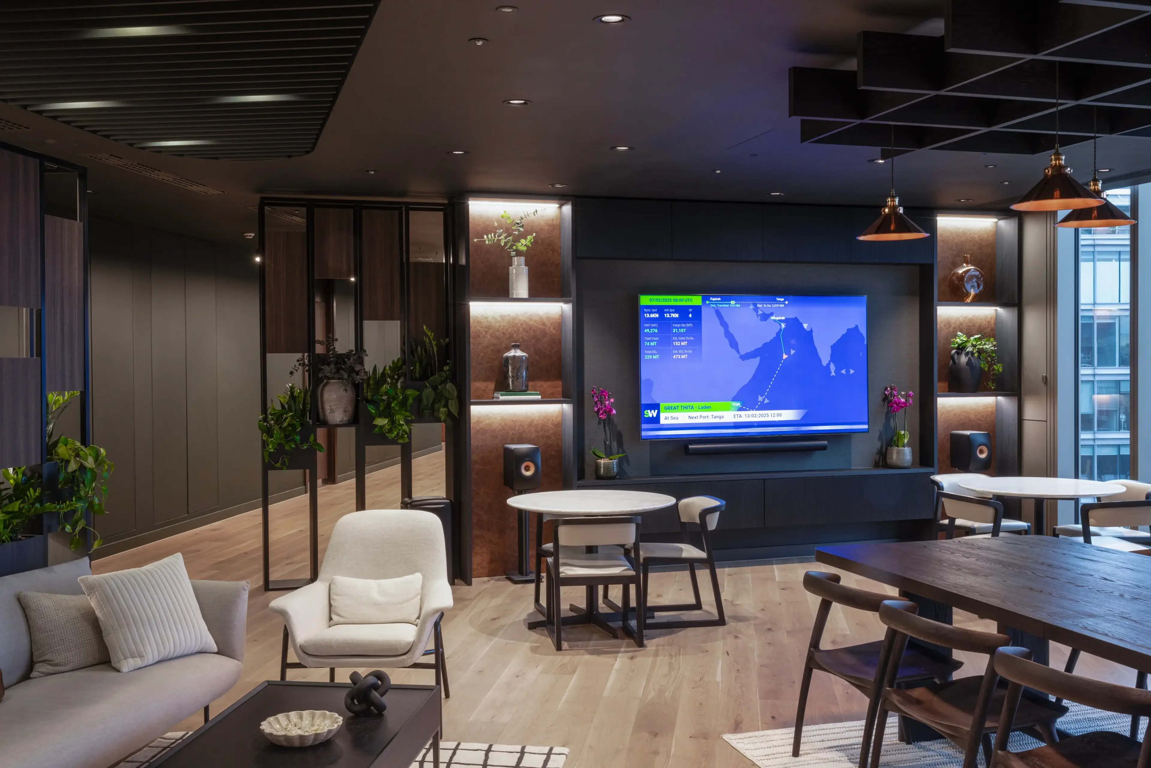 A stylish office lounge with a large display screen on a feature wall, surrounded by illuminated shelving with decorative objects and plants. The space includes a mix of soft seating, round tables, and a dark wooden dining table.