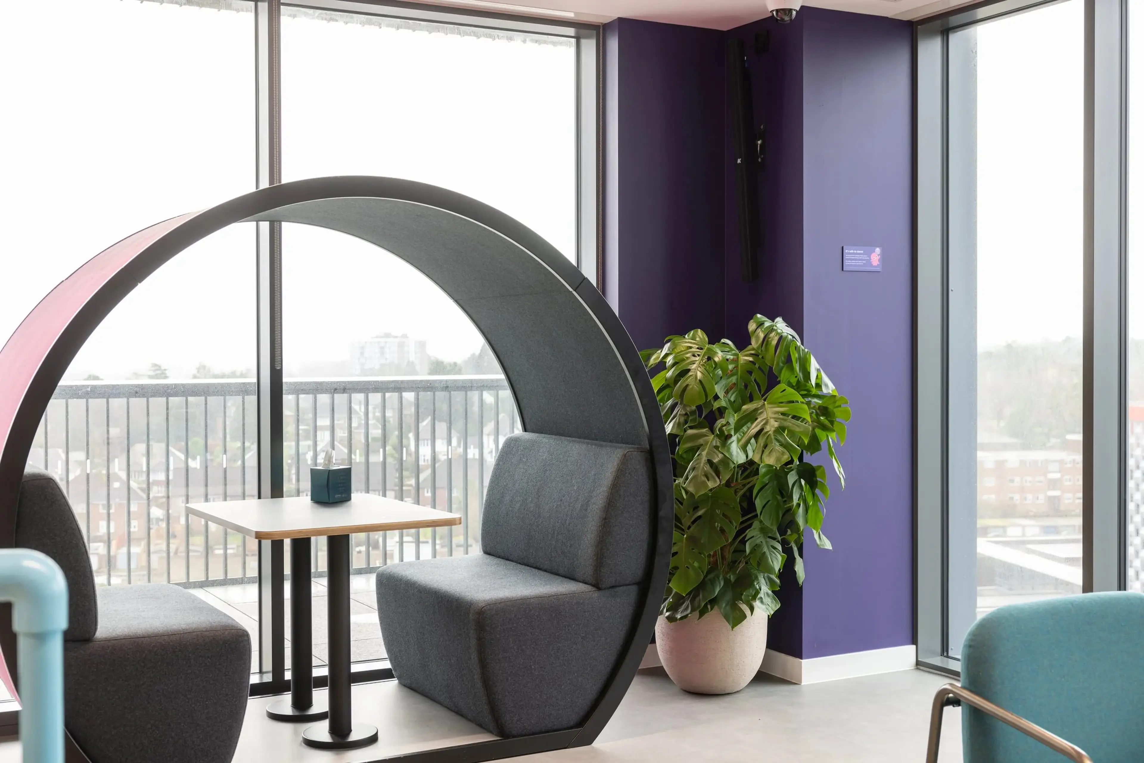 Modern office space with circular seating booth, small table, and large potted plant by floor-to-ceiling windows overlooking a cityscape.