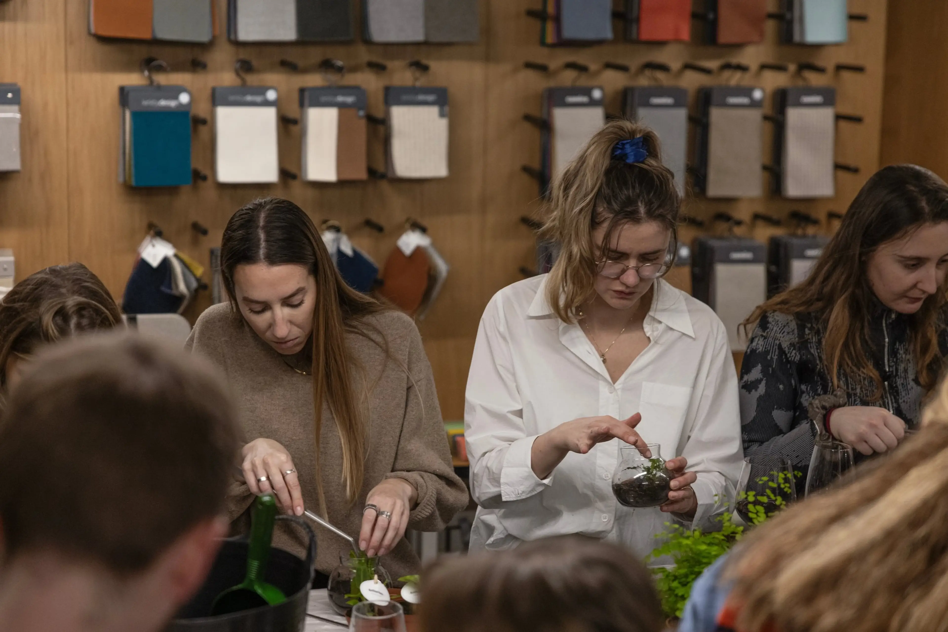 People engaged in a planting activity, preparing terrariums with soil. Fabric samples hang on the wall in the background.