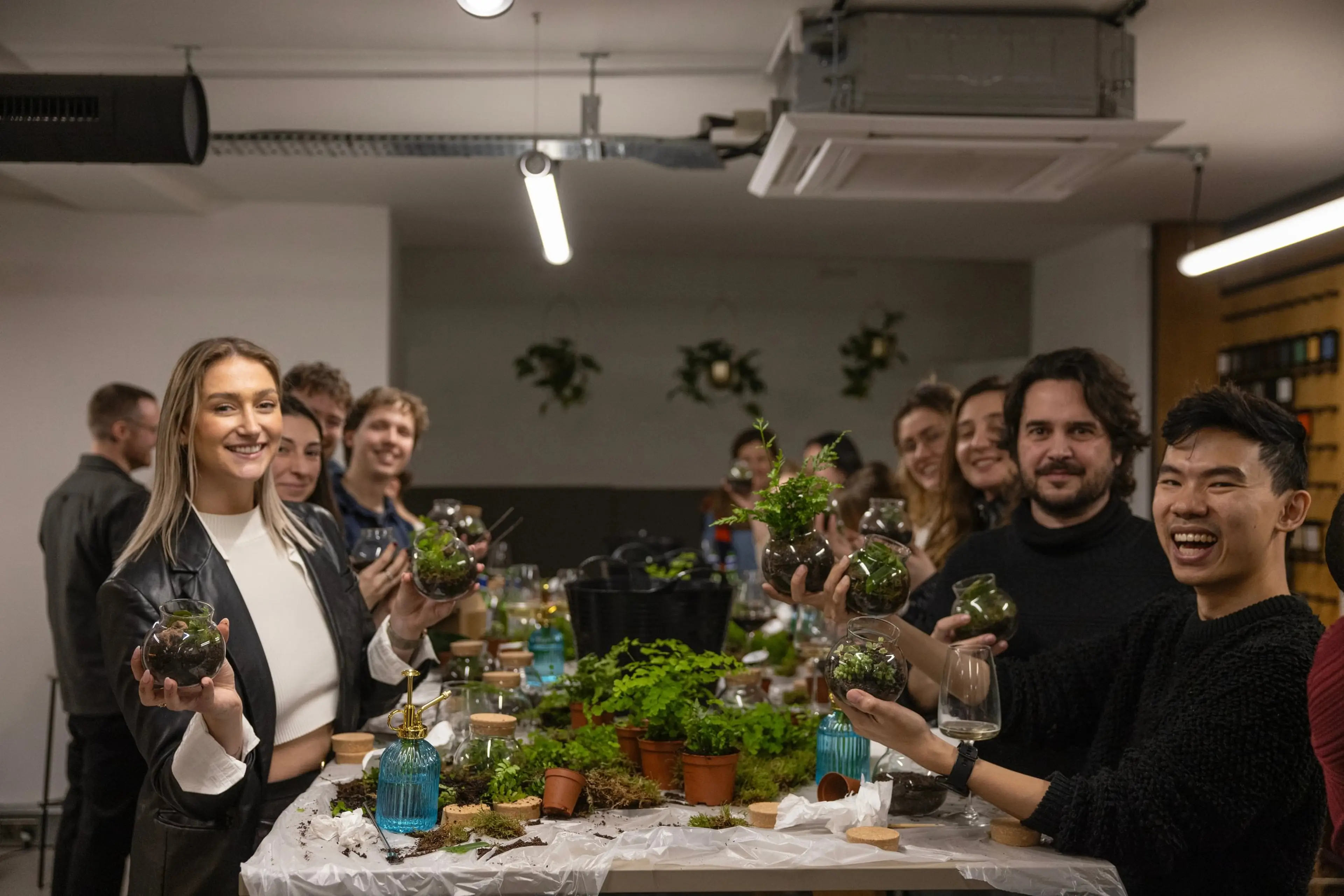 A group of people smiling and holding small terrariums in a workshop setting, with plants and tools on the table.