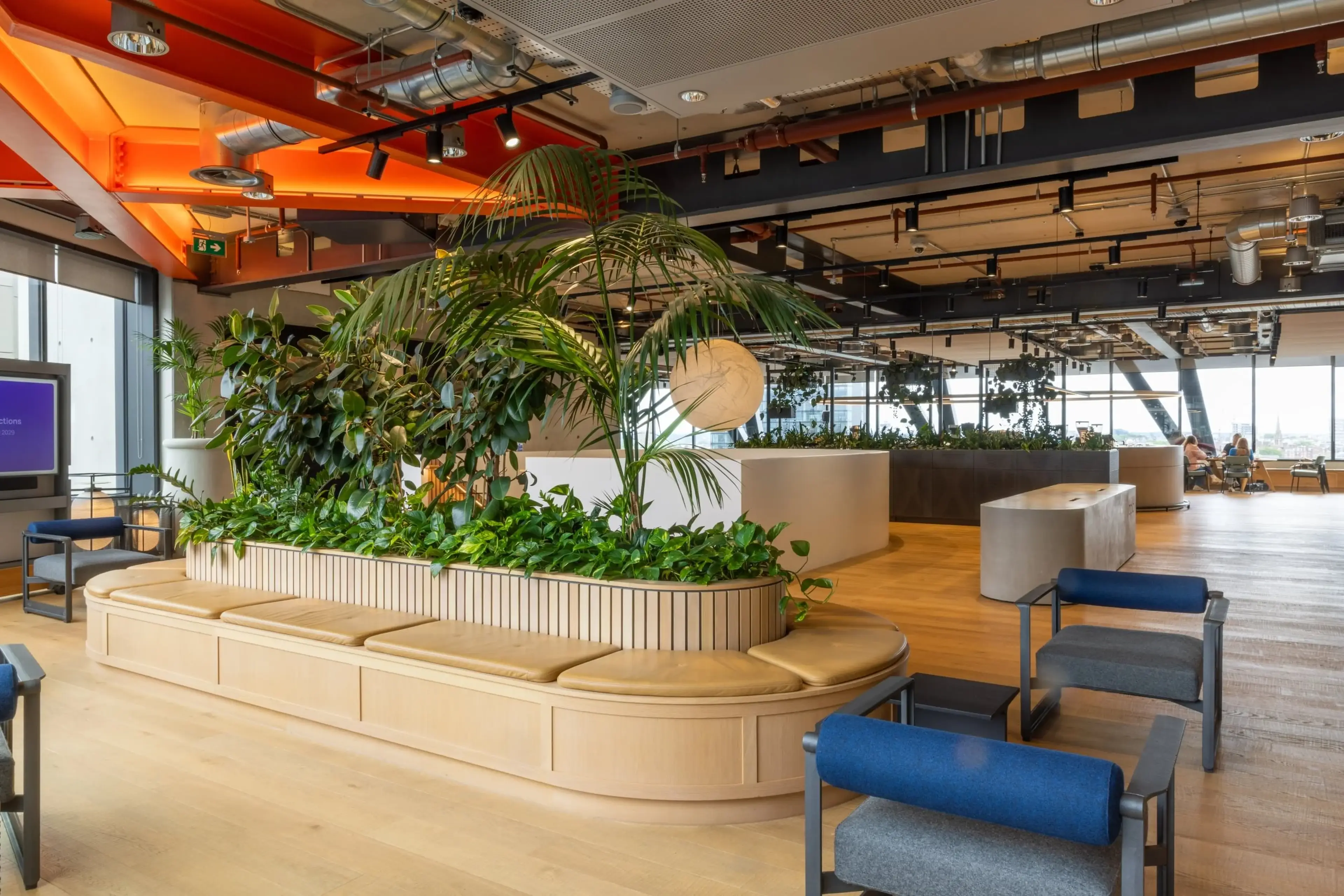 Modern office space with wooden flooring, indoor plants, cushioned seating, and exposed ceiling. Large windows offer natural light.