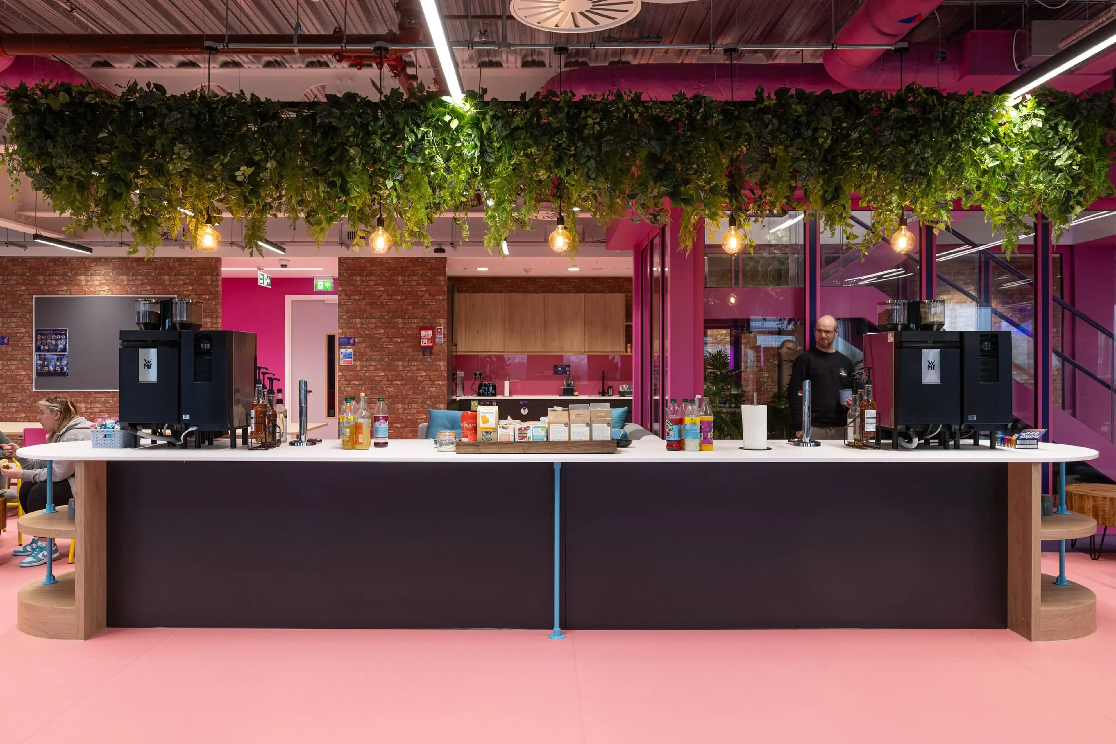 Modern office kitchen with a long counter, coffee machines, hanging plants, and a person preparing drinks. Bright pink walls and ceiling details.
