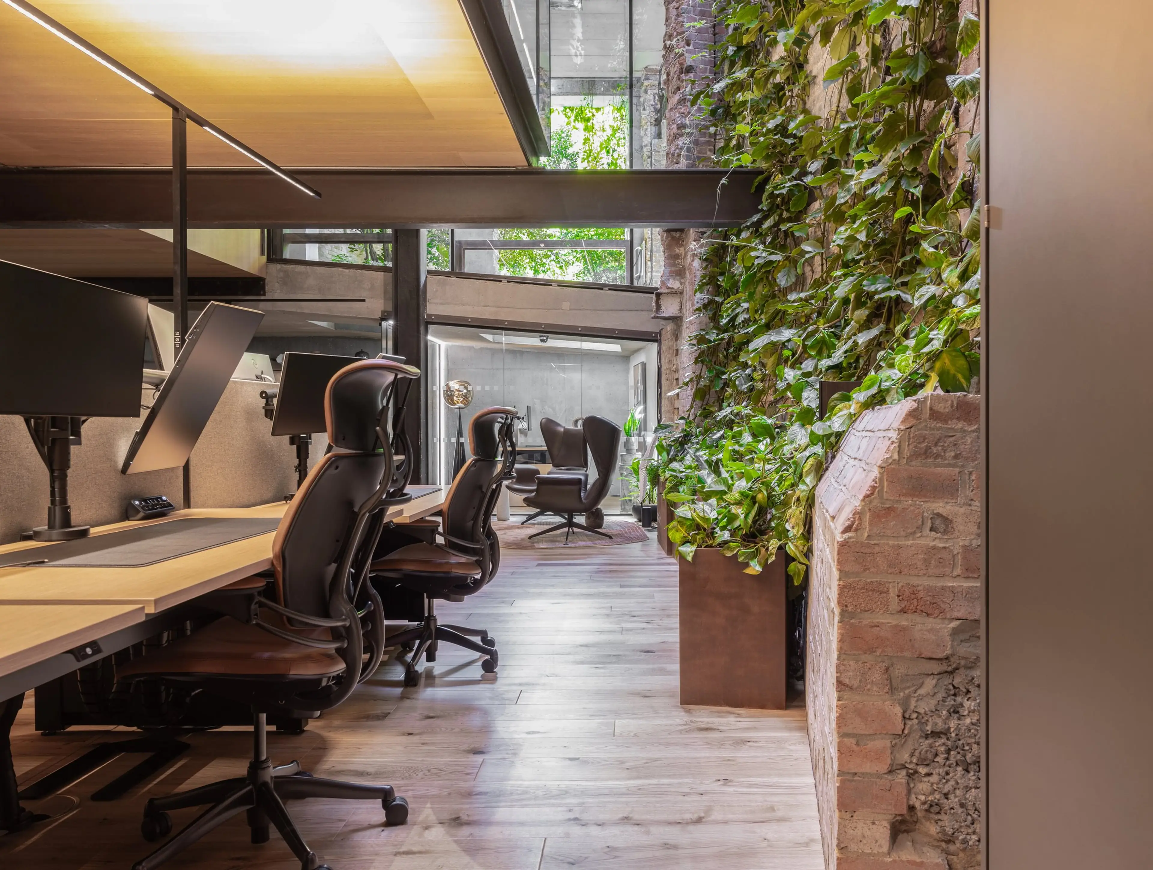 Modern office with ergonomic chairs, wooden desks, and a vertical green wall. Large windows provide natural light.