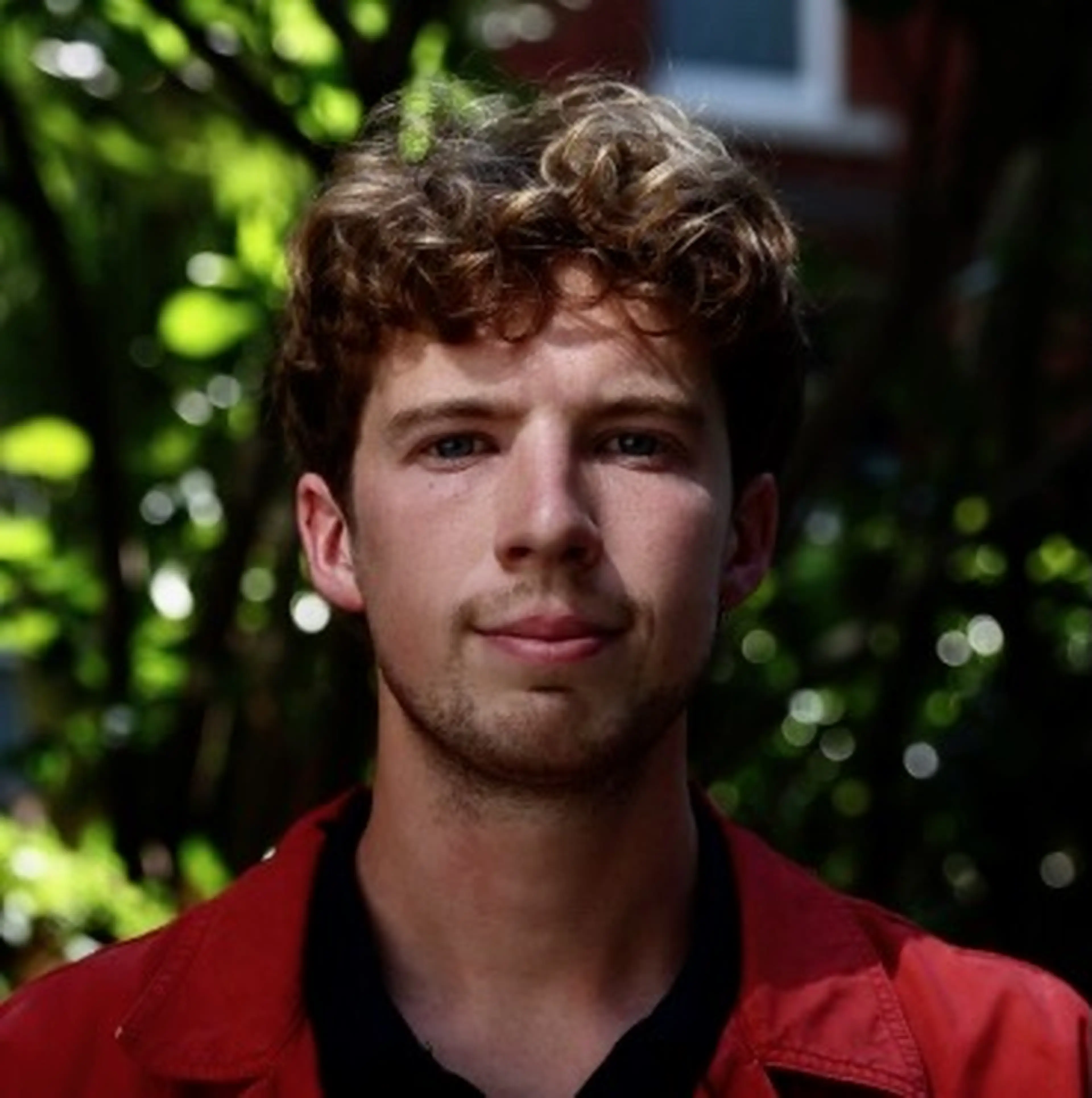 A person with curly hair wearing a red jacket stands outdoors, with greenery and dappled sunlight in the background.