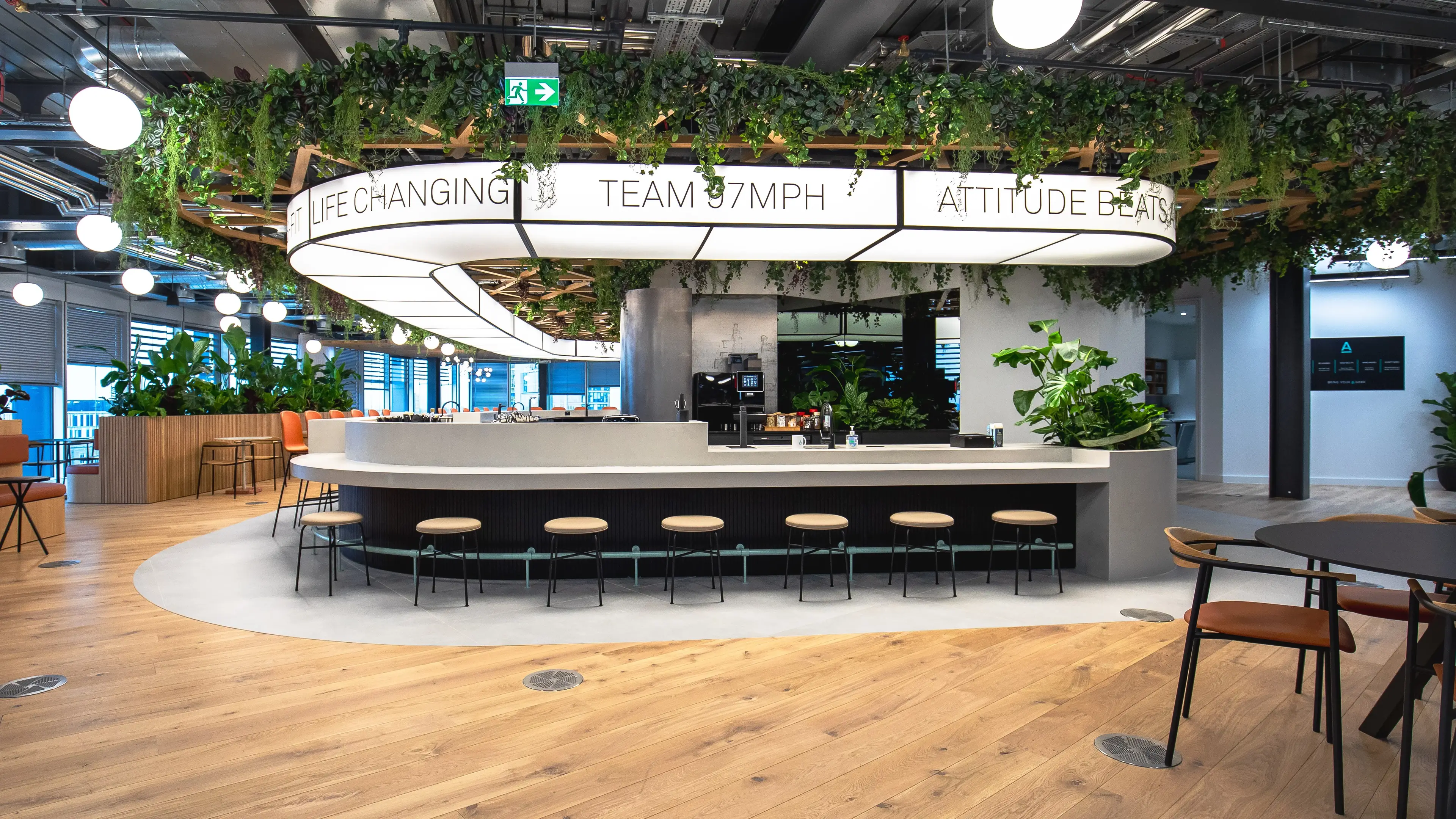 Modern office break area with a circular bar, stools, hanging plants, and motivational signs. Bright lighting and wooden flooring.