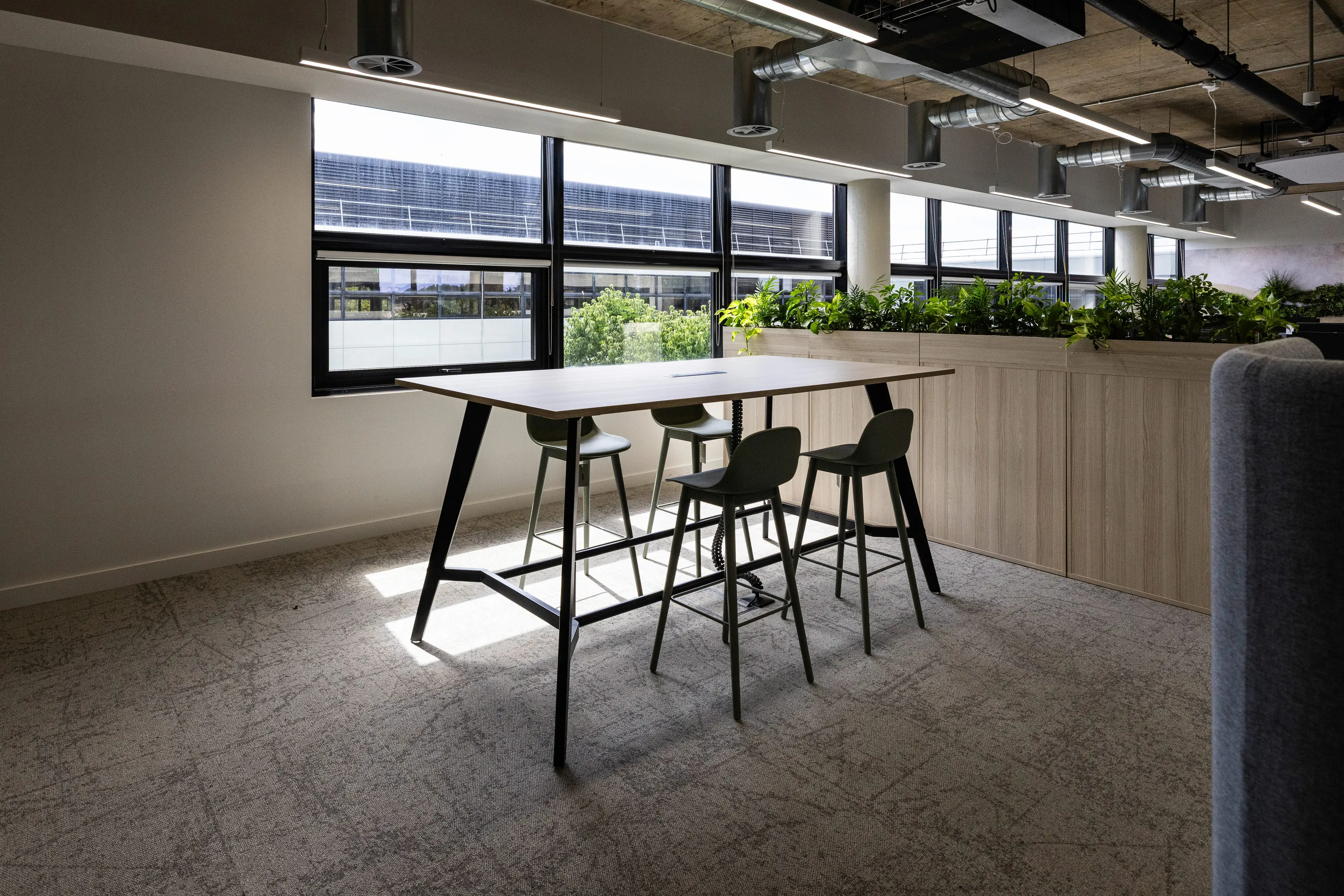 Modern office space with a high table, four green stools, large windows, and a planter filled with greenery, under exposed ductwork.