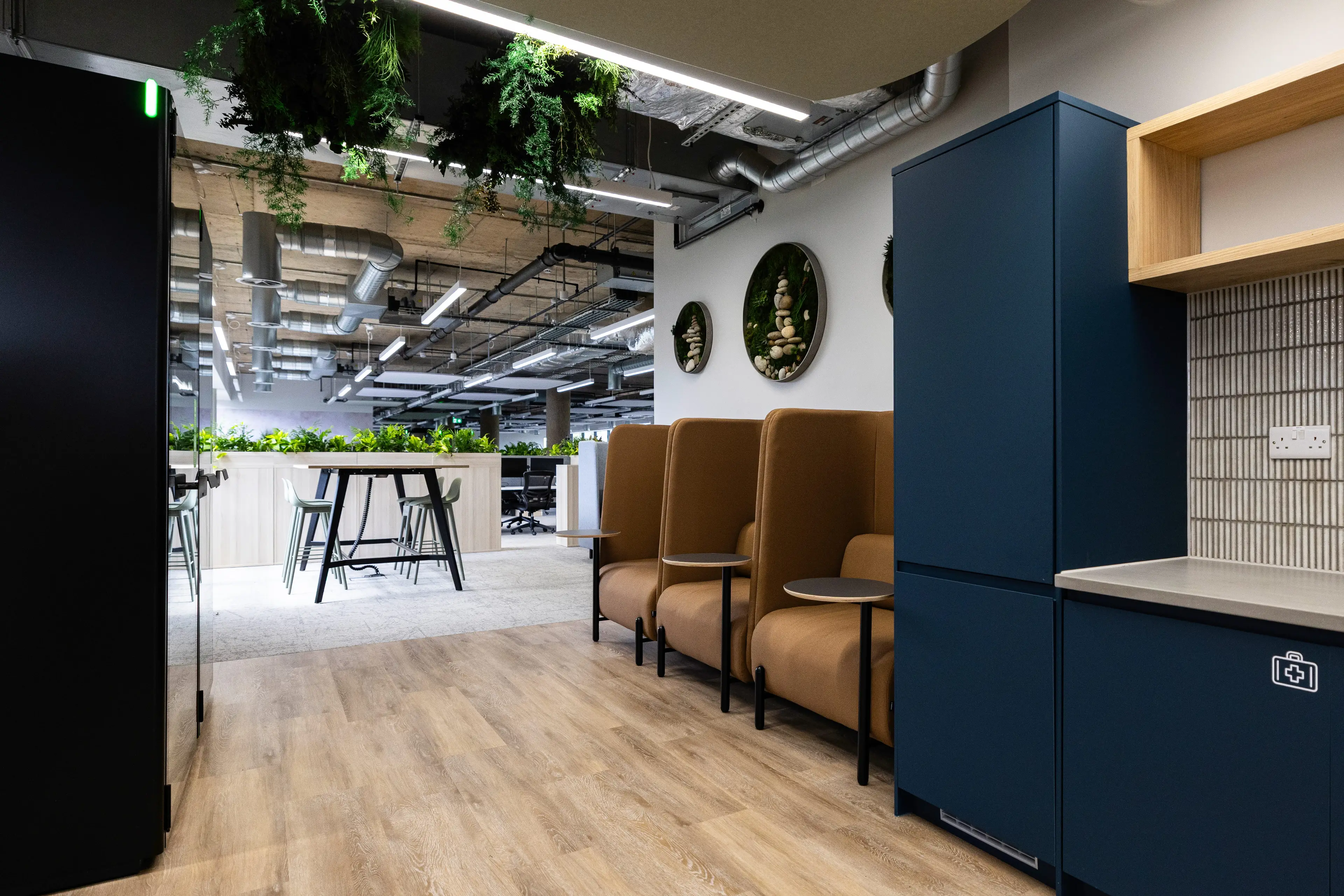 Modern office space with blue cabinetry, brown seating, overhead plants, and a bar-height table with chairs, featuring exposed ductwork.