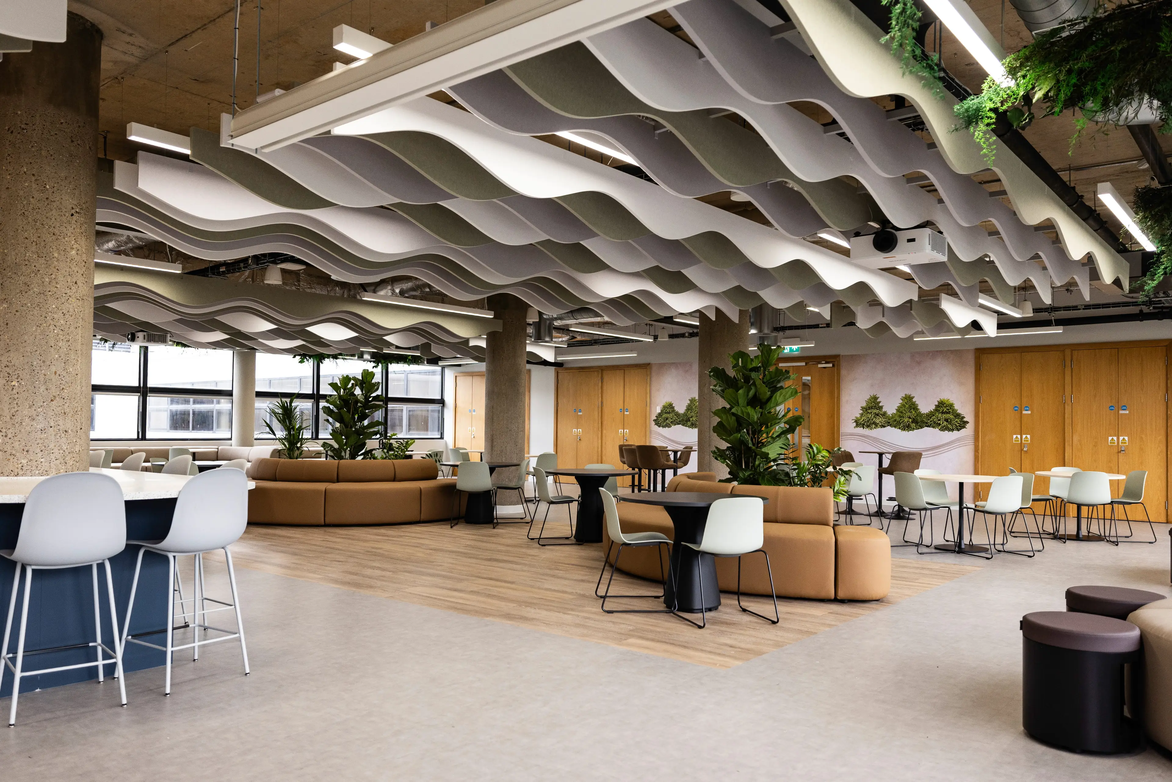 Modern open office space with wave-like ceiling panels, plants, brown couches, and white chairs around tables on wooden flooring.