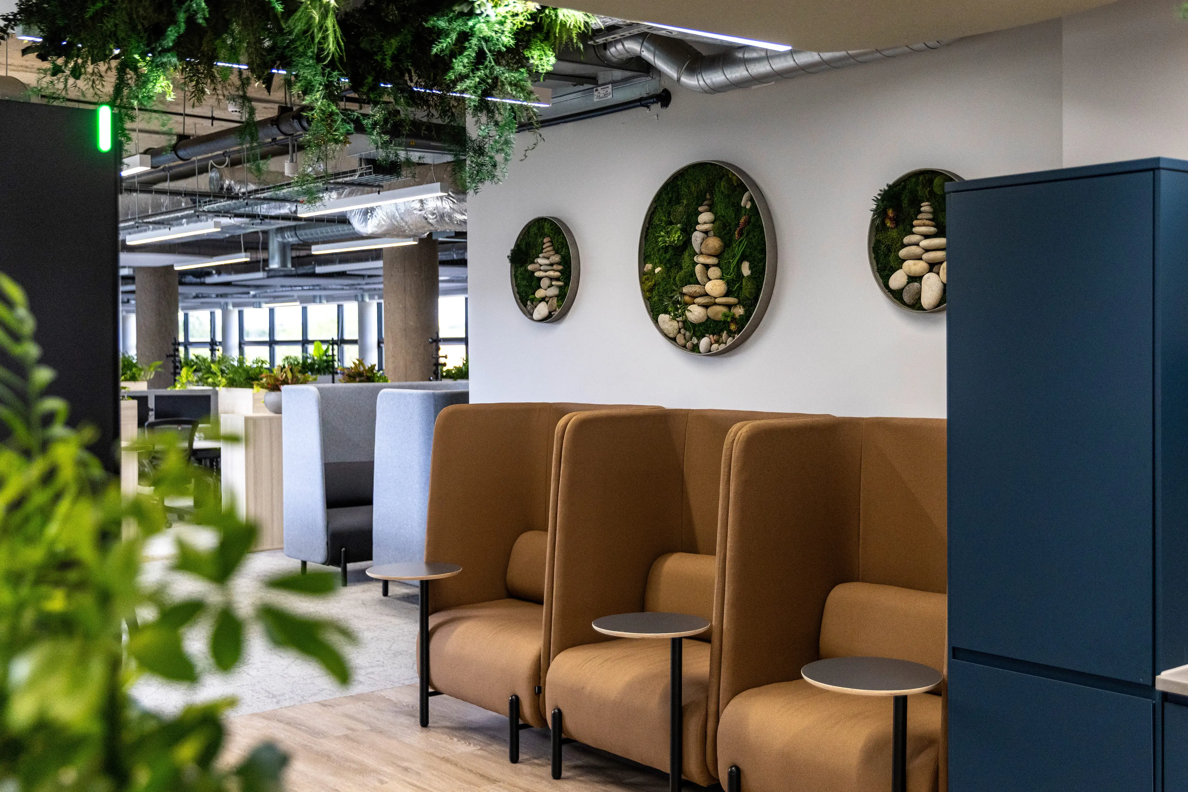 Modern office lounge with brown high-back chairs, round tables, wall art featuring stones and greenery, and overhead greenery hanging from the ceiling.