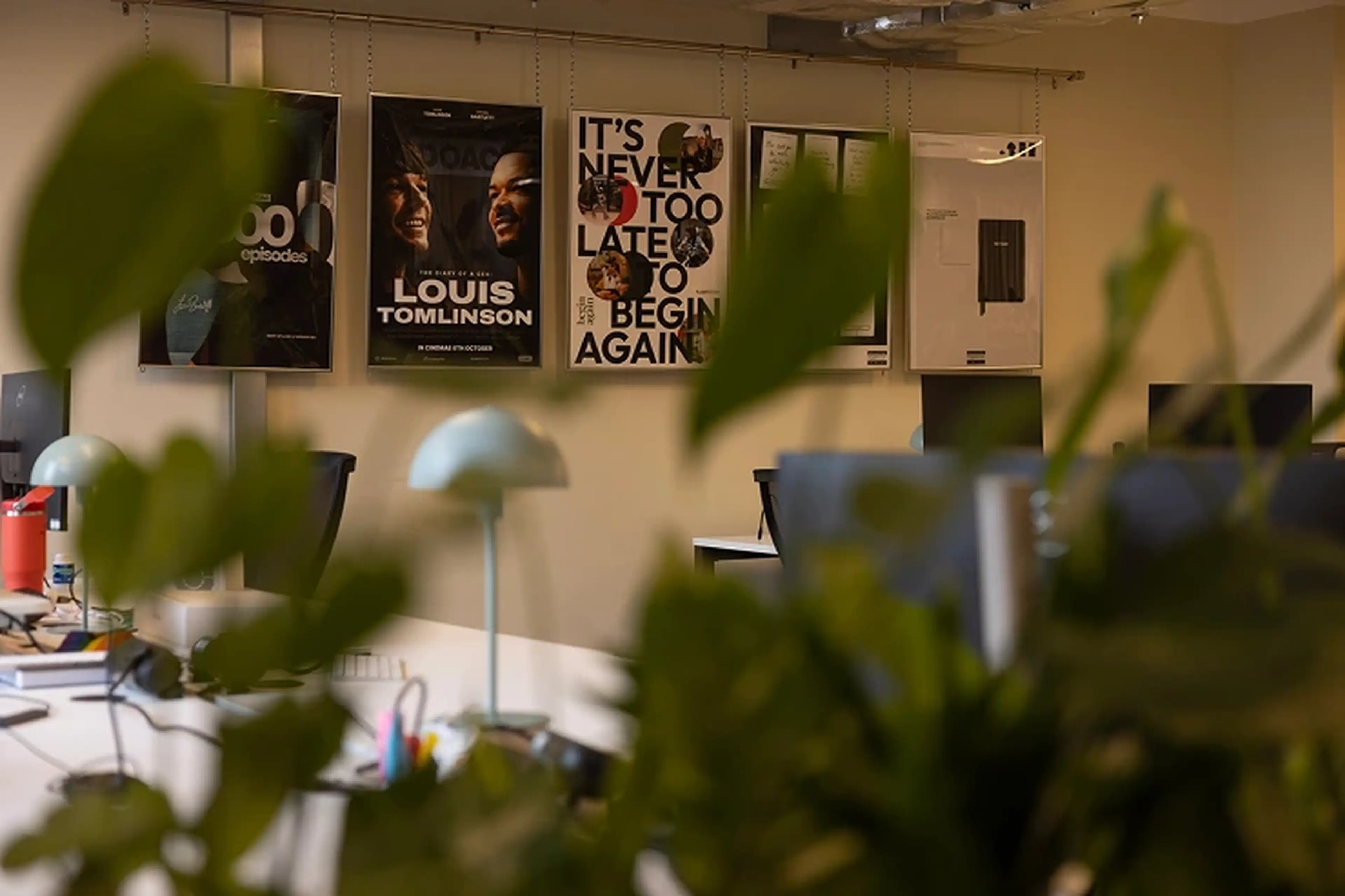 Office space with desks, lamps, and computers, decorated with motivational posters on the wall. Foreground features out-of-focus green plants.