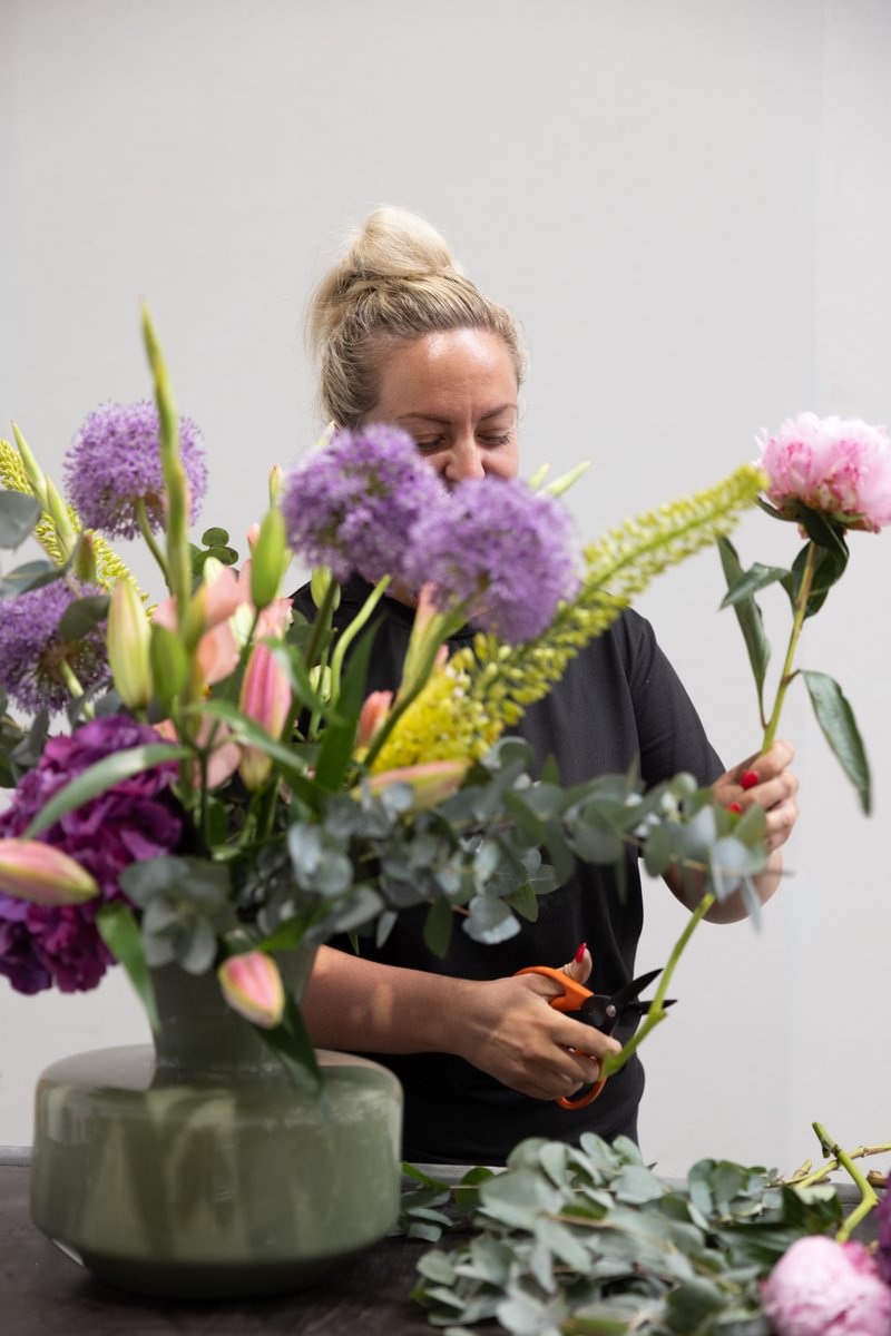 A woman arranging a colorful bouquet of flowers, including lilies and alliums, holds a stem and scissors in her hands, smiling happily.