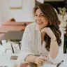 Caitriona Hughes, Creative Director at Bright Projects, smiling while working at a design table with material samples and a pen in hand. She is wearing a white textured blouse and gold jewellery, with a warm and inviting interior in the background.