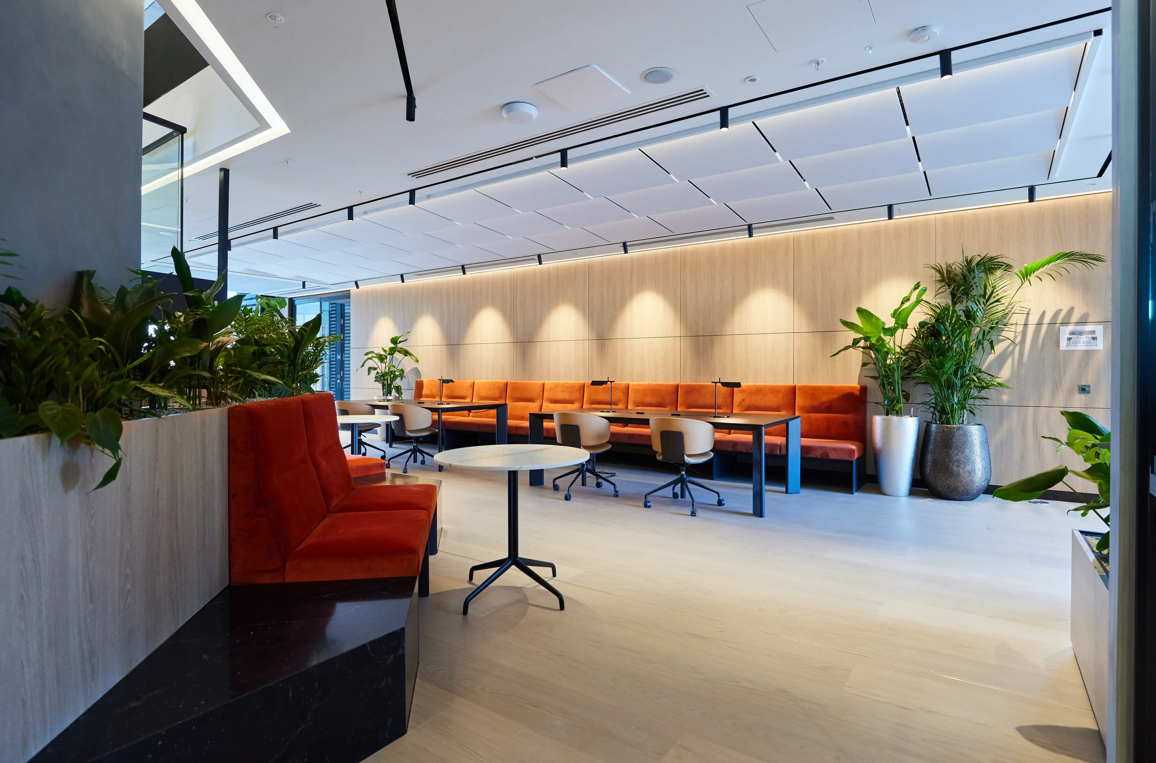 Modern office lounge with orange sofas, round tables, swivel chairs, and large potted plants under ceiling lights and wooden paneled walls.