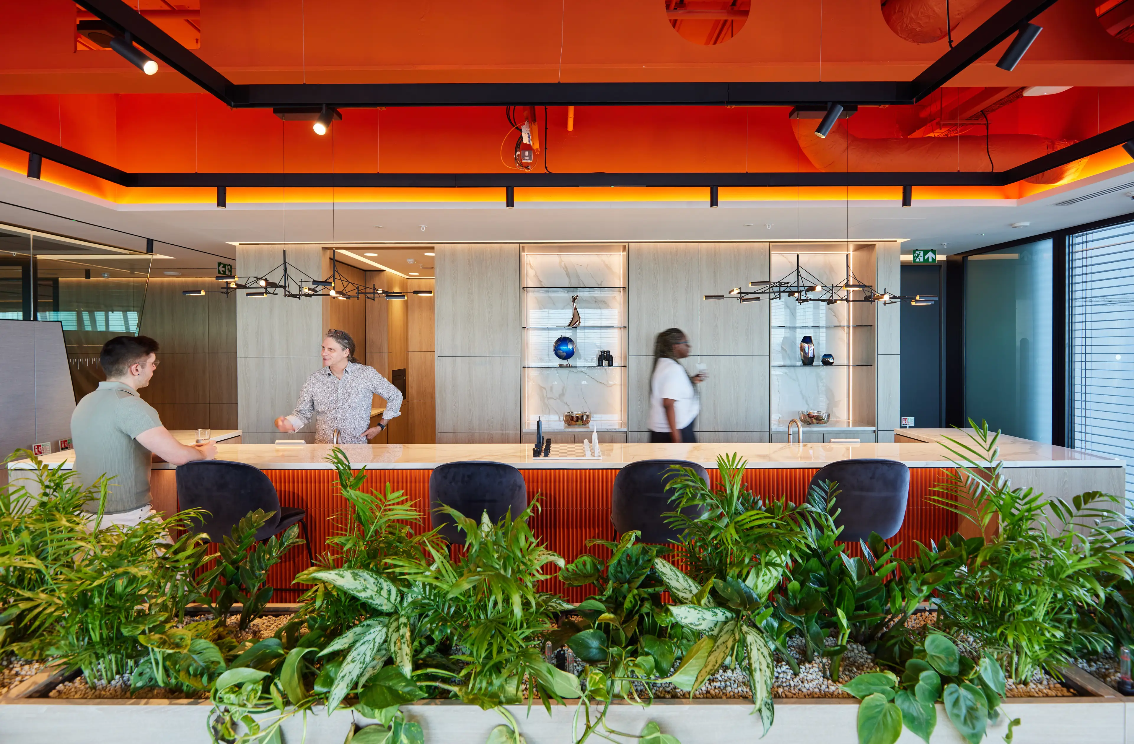 Modern office lounge with two people conversing at a counter, surrounded by greenery; a third person walks by. Warm lighting and decor.