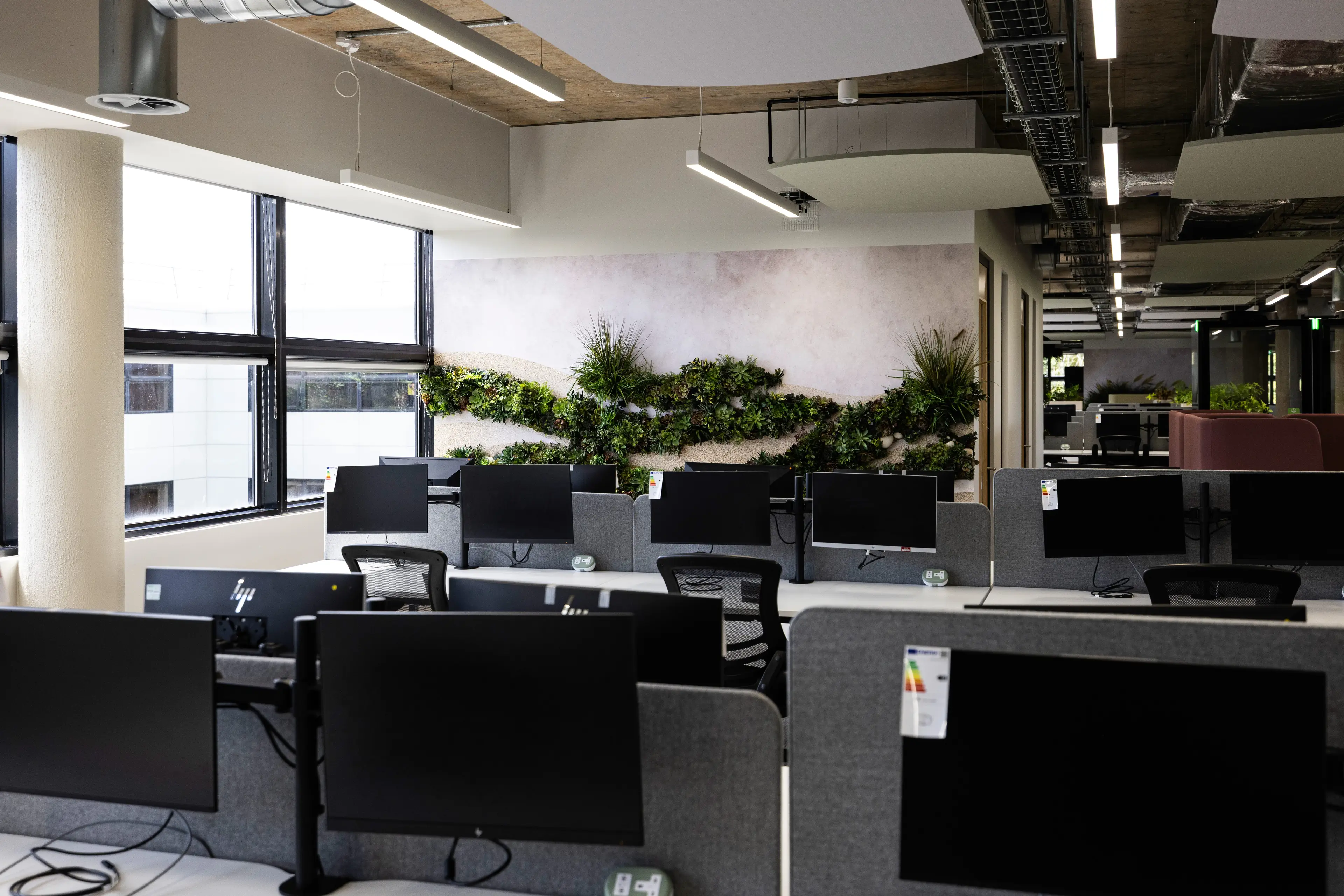 Modern office space with multiple desks, monitors, and a green plant wall, featuring large windows and exposed ceiling design.