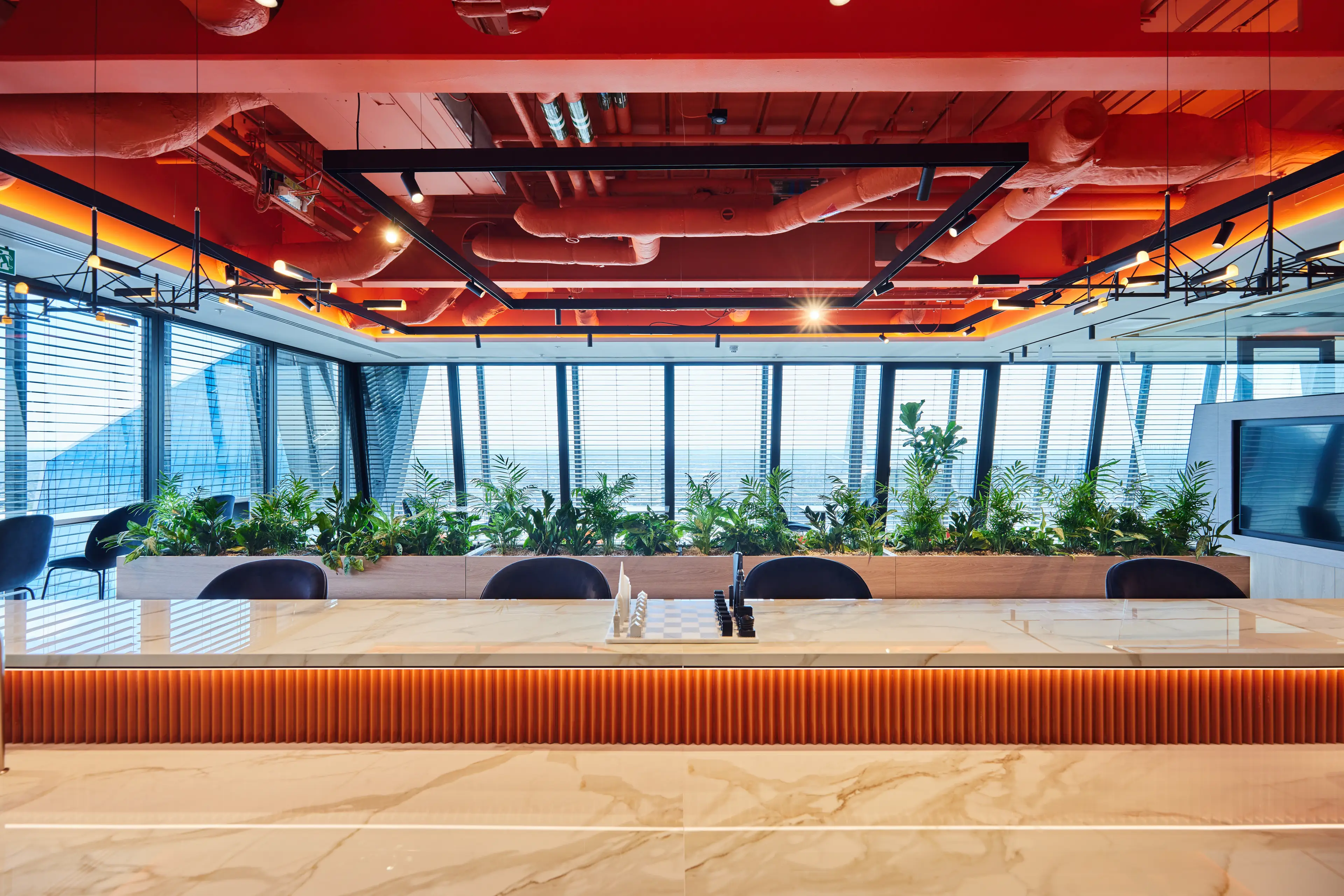 Modern office space with a marble countertop, red ceiling, industrial piping, hanging lights, large windows, and indoor plants.
