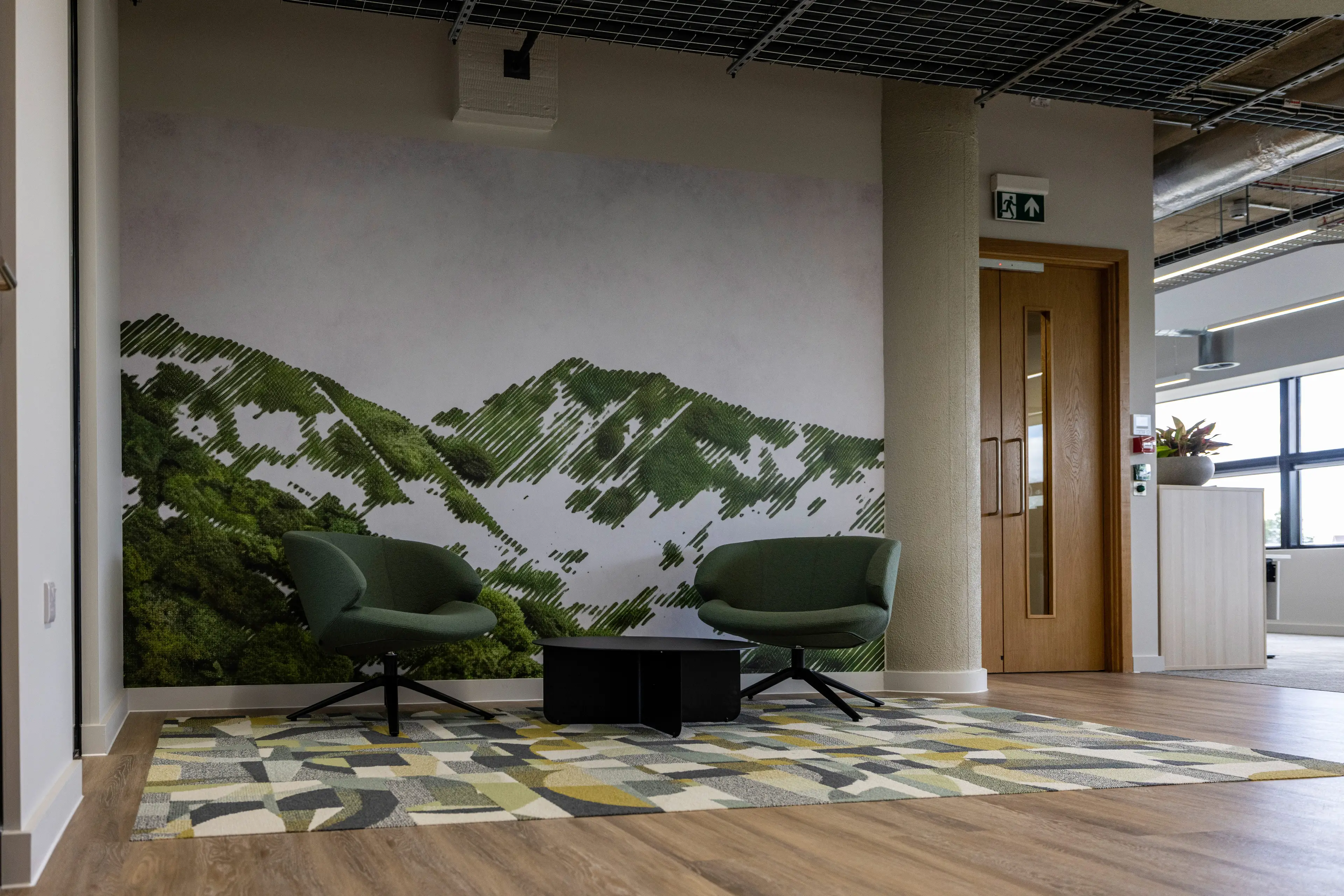 Office area with two green armchairs and a small table on a patterned rug, set against a mural depicting a stylized mountain landscape.