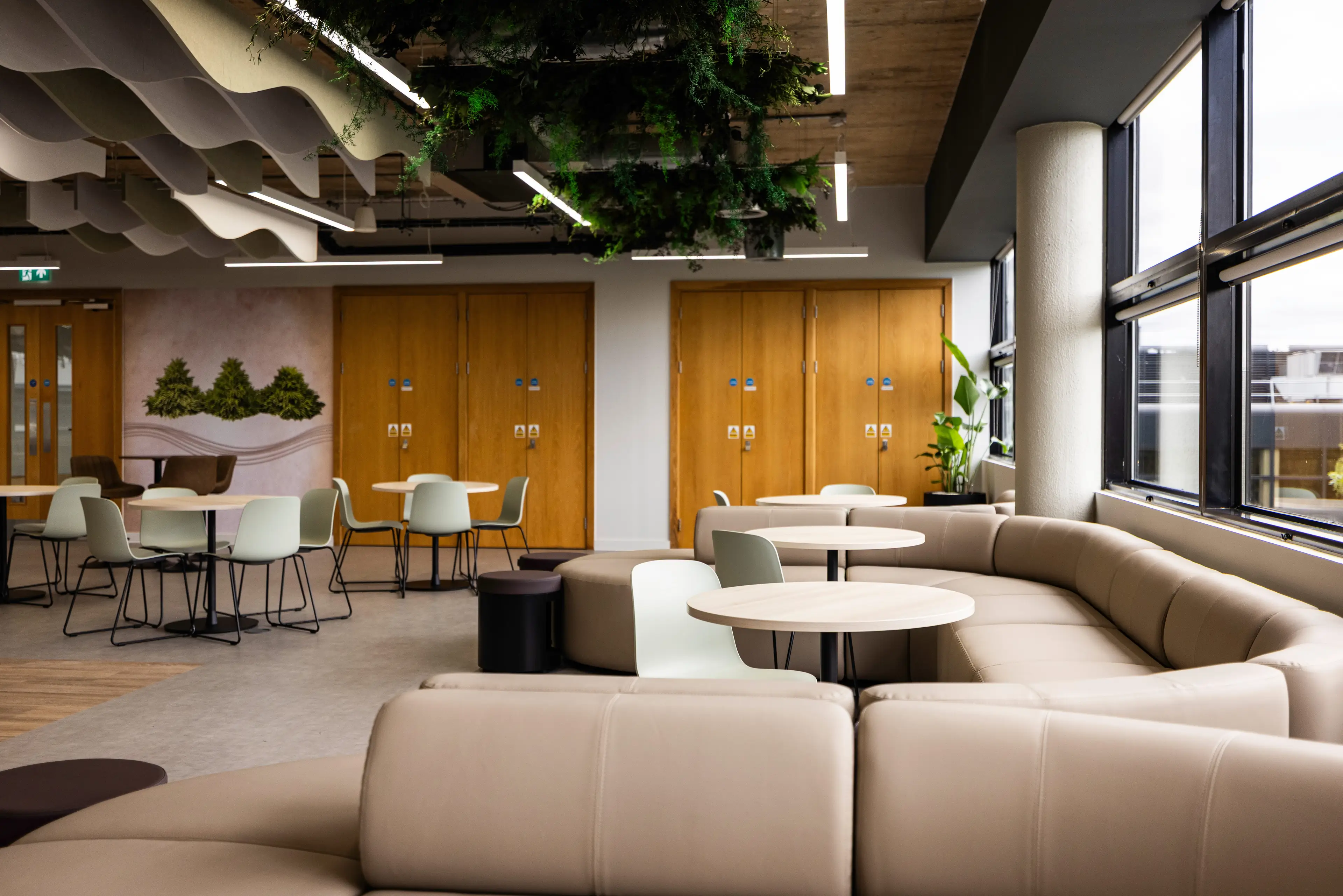Modern Office lounge area with beige couches, round tables, and chairs. Ceiling has wave-like panels and greenery. Large windows on the right.