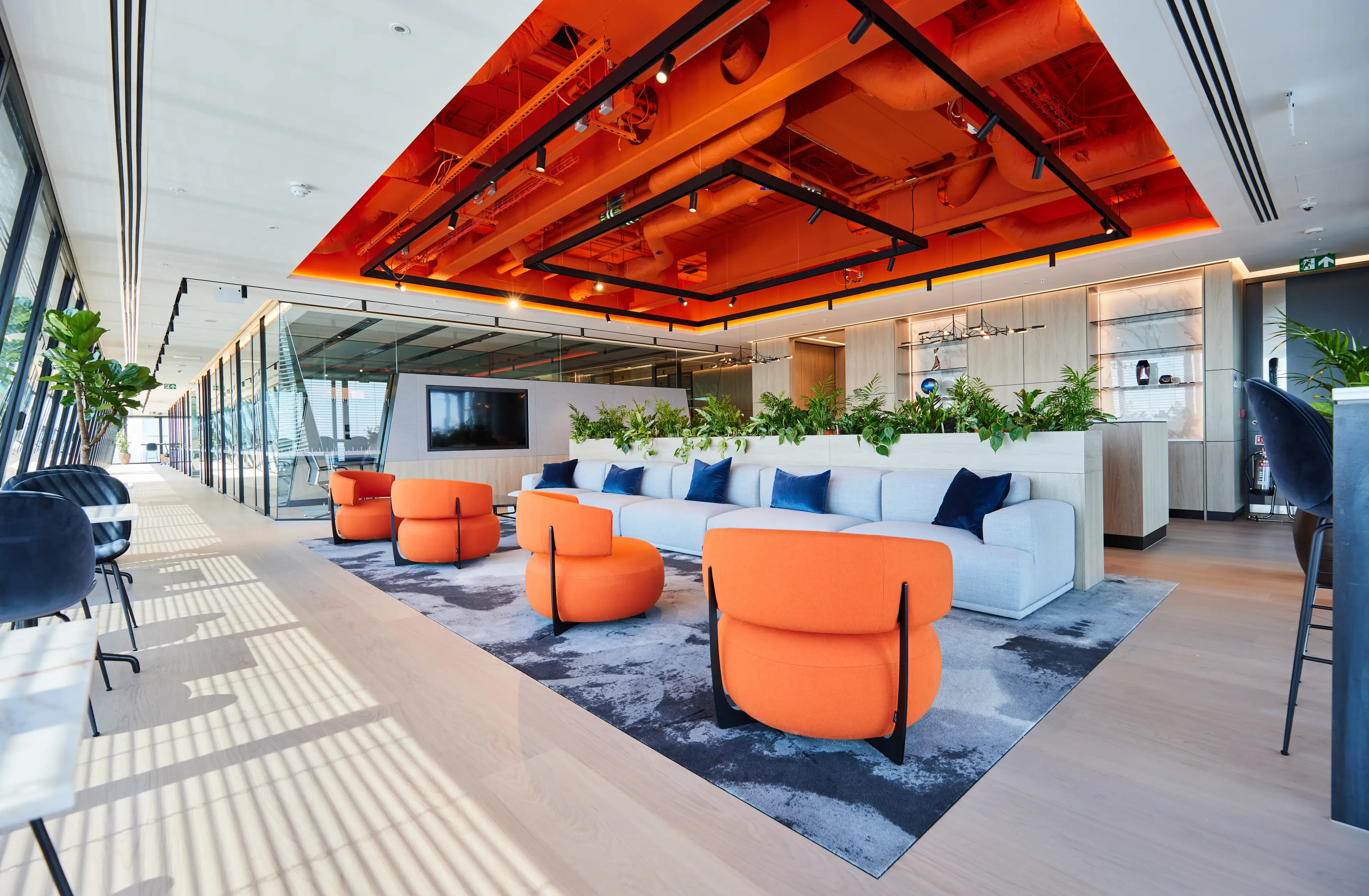 Modern office lounge with orange ceiling, vibrant orange chairs, a grey sofa with blue cushions, plants, and large windows with natural light.