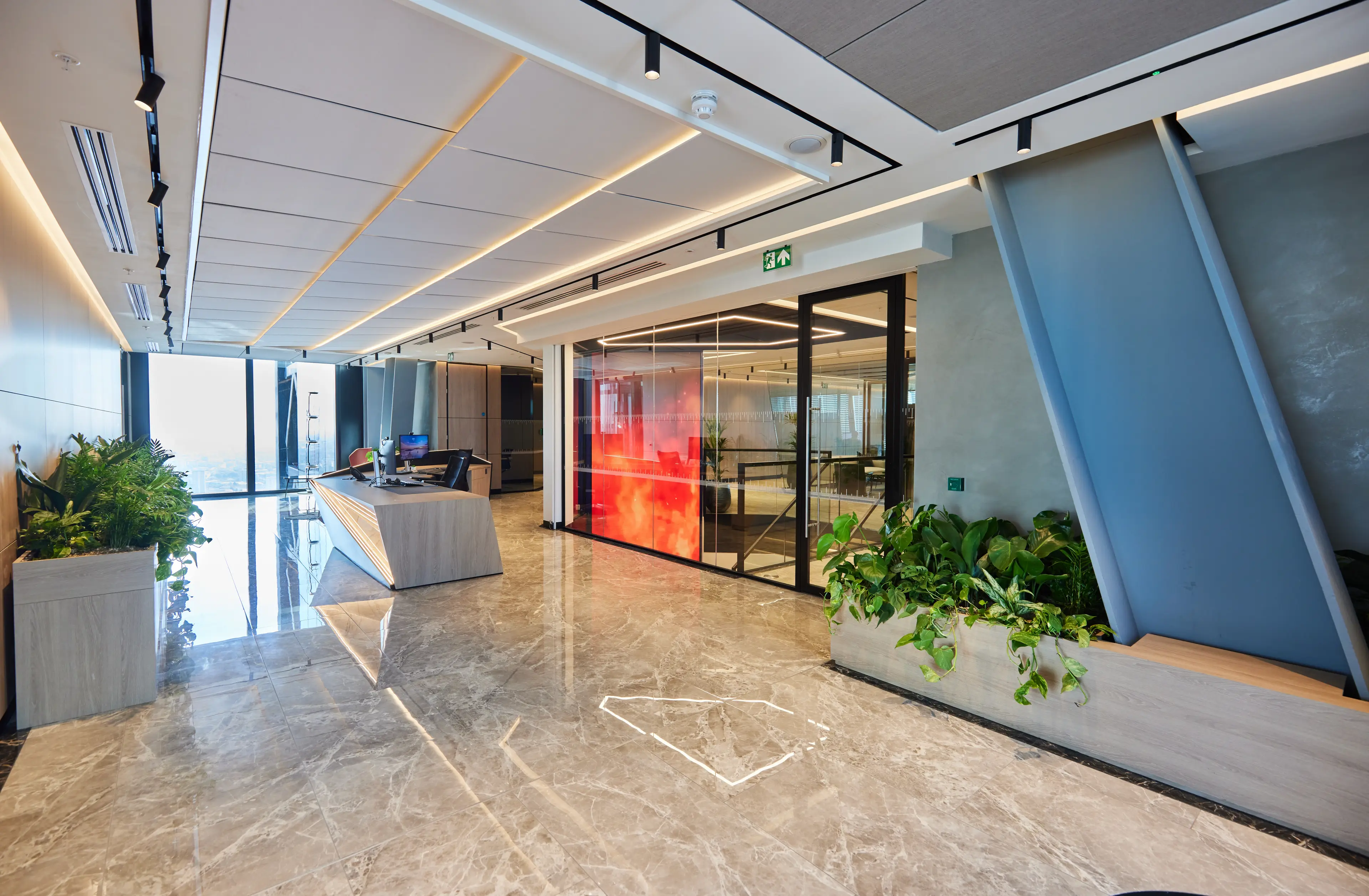 Modern office lobby with marble flooring, a sleek reception desk, large windows, and indoor plants. A glass partition displays a vibrant design.