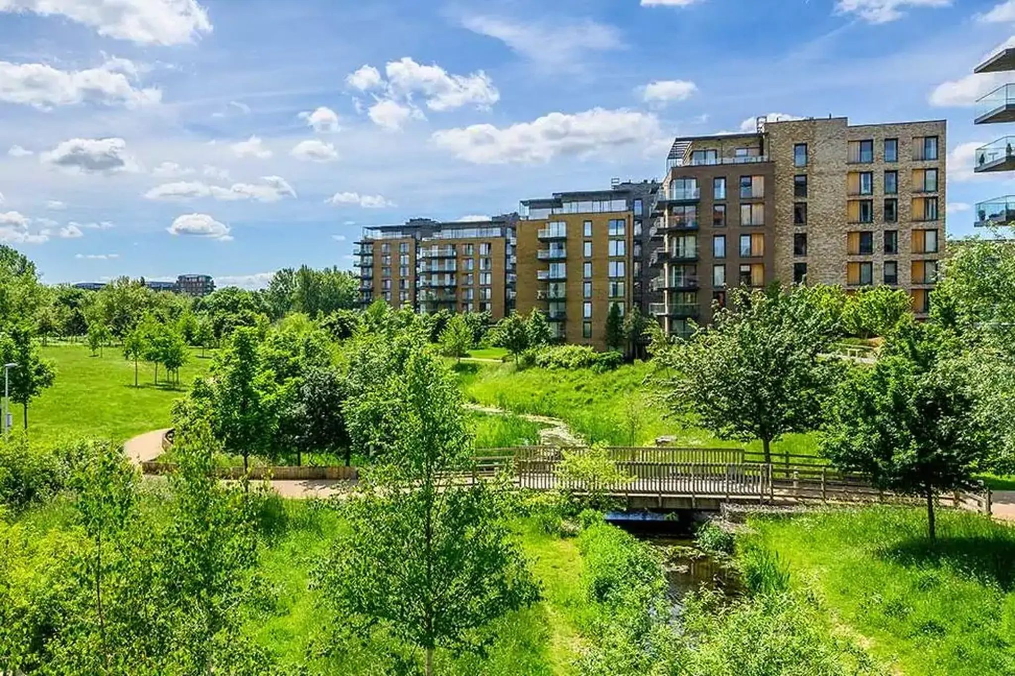 Kidbrooke Village: A sustainable urban development featuring modern apartment buildings surrounded by green spaces, trees, and a natural waterway with a wooden footbridge. Designed with biodiversity net gain (BNG) in mind, the landscape integrates ecological enhancements, promoting habitat creation and urban greenery.
