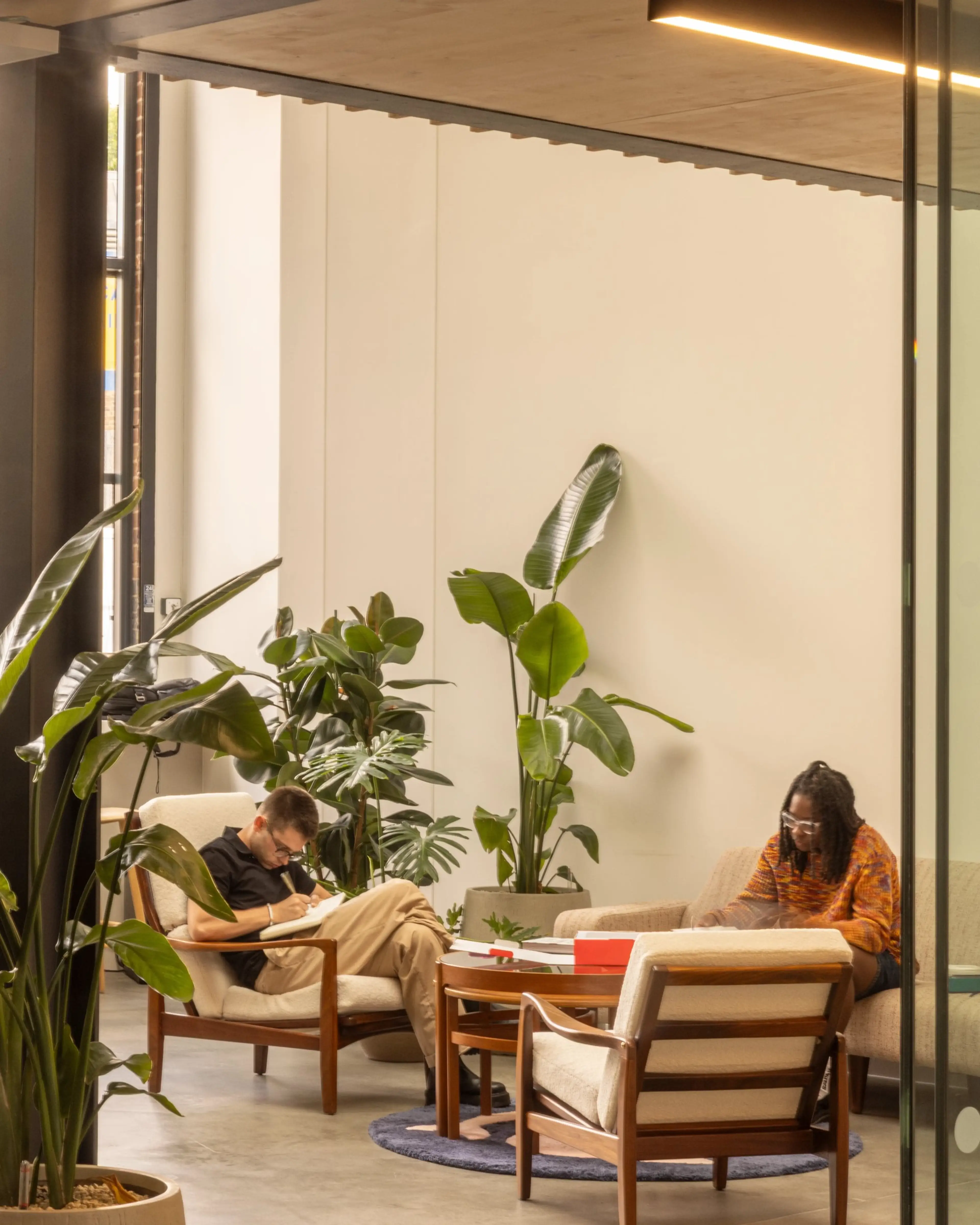 A communal workspace in a bright lit office, with two people sat reading books. There are office plants in the foreground and background.