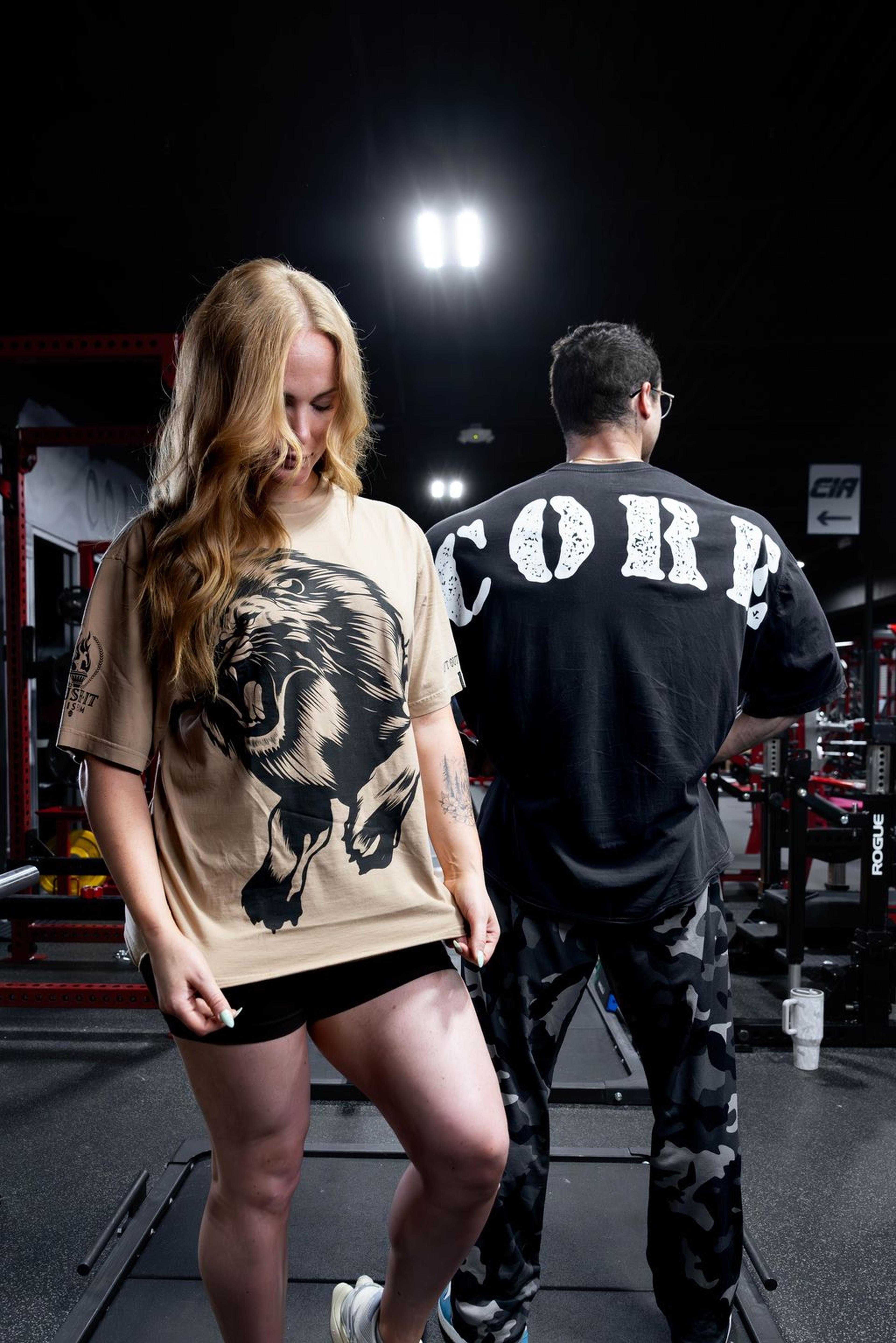 Two people in a gym, one facing forward wearing a lion print shirt, the other facing away wearing a shirt with "CORP" text.