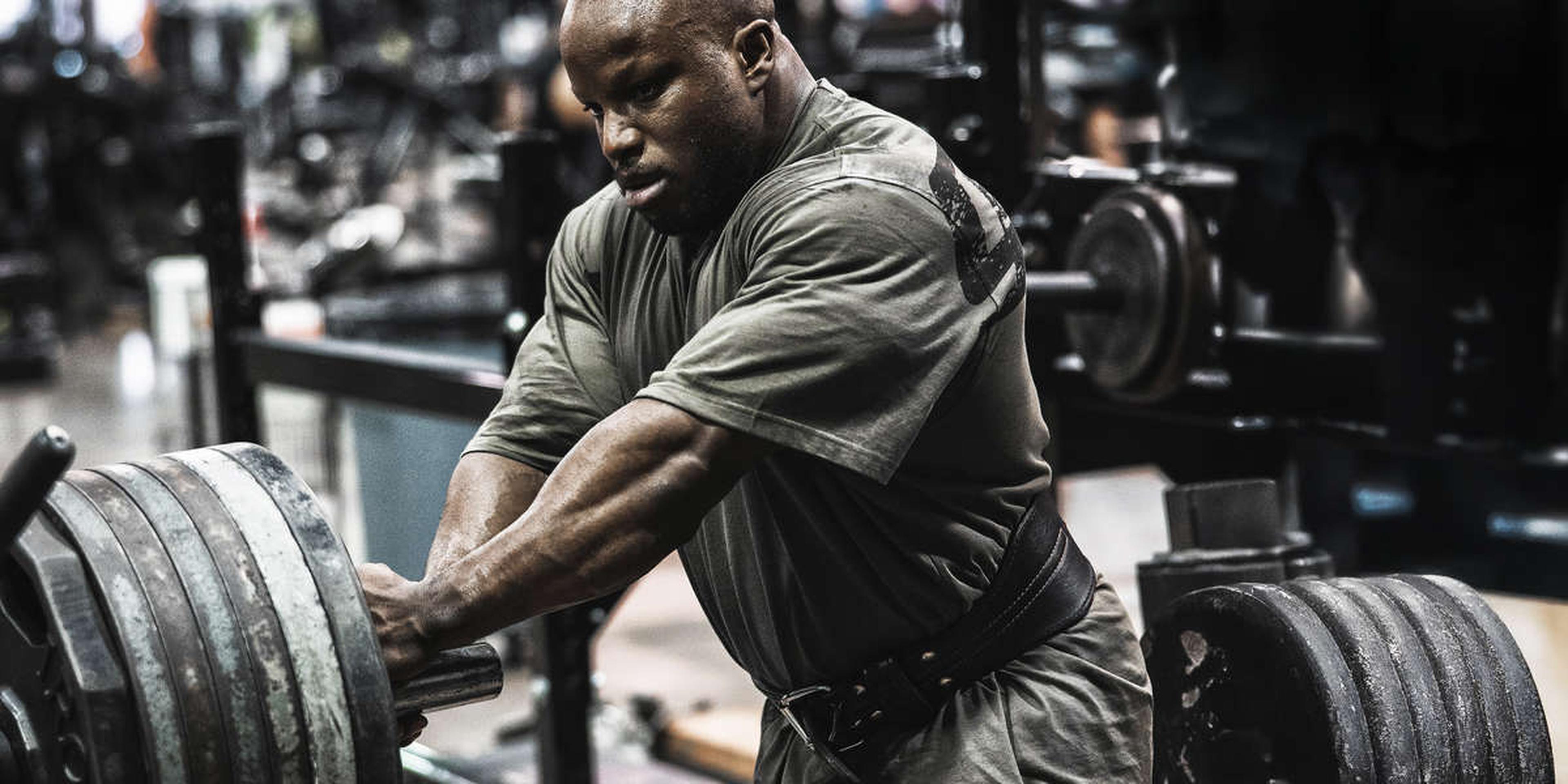 A man in a gym prepares to lift a heavily loaded barbell, wearing a weightlifting belt and a gray shirt.
