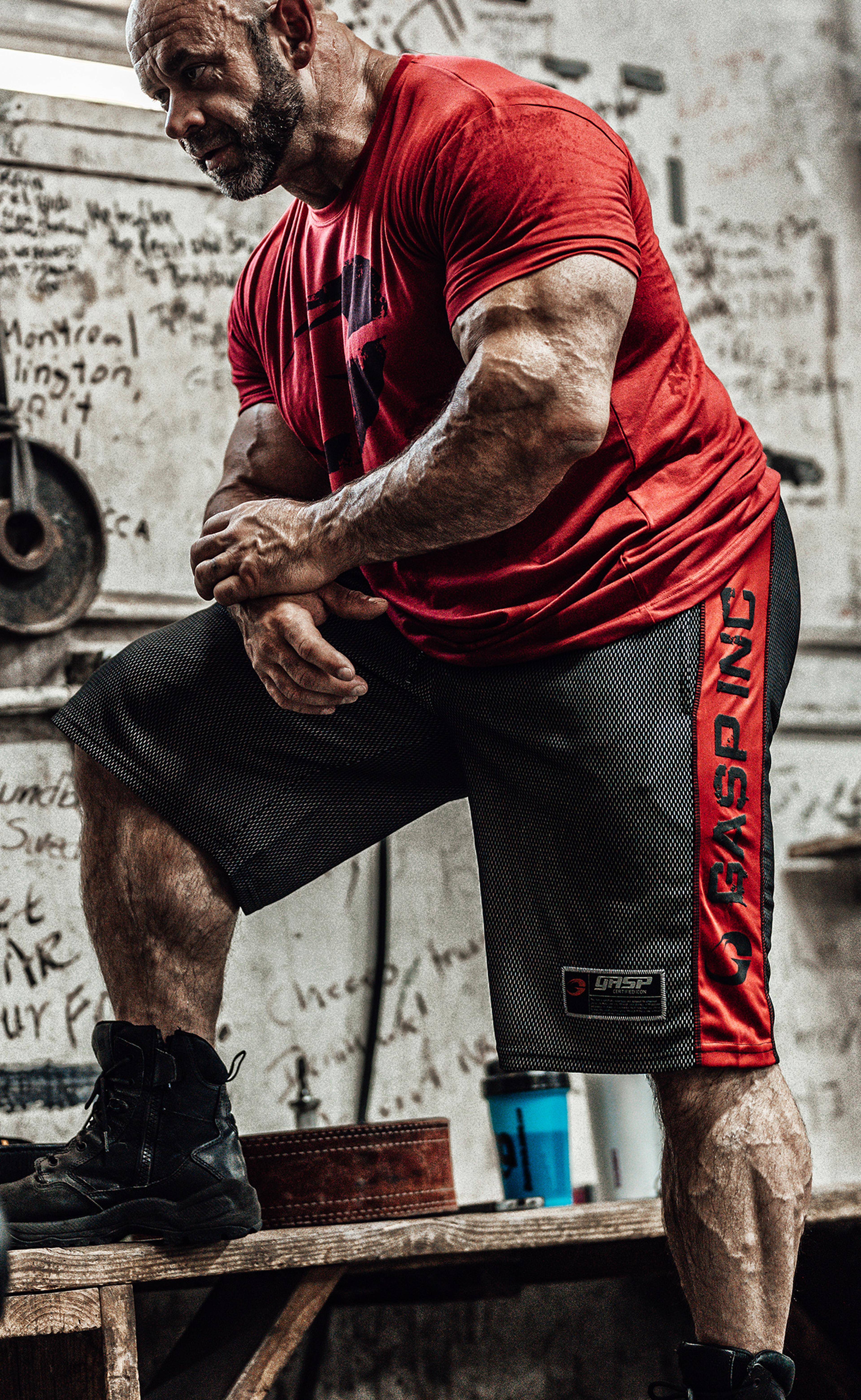 Muscular man in a red shirt and black shorts, flexing his arms in a gym with graffiti-covered walls.