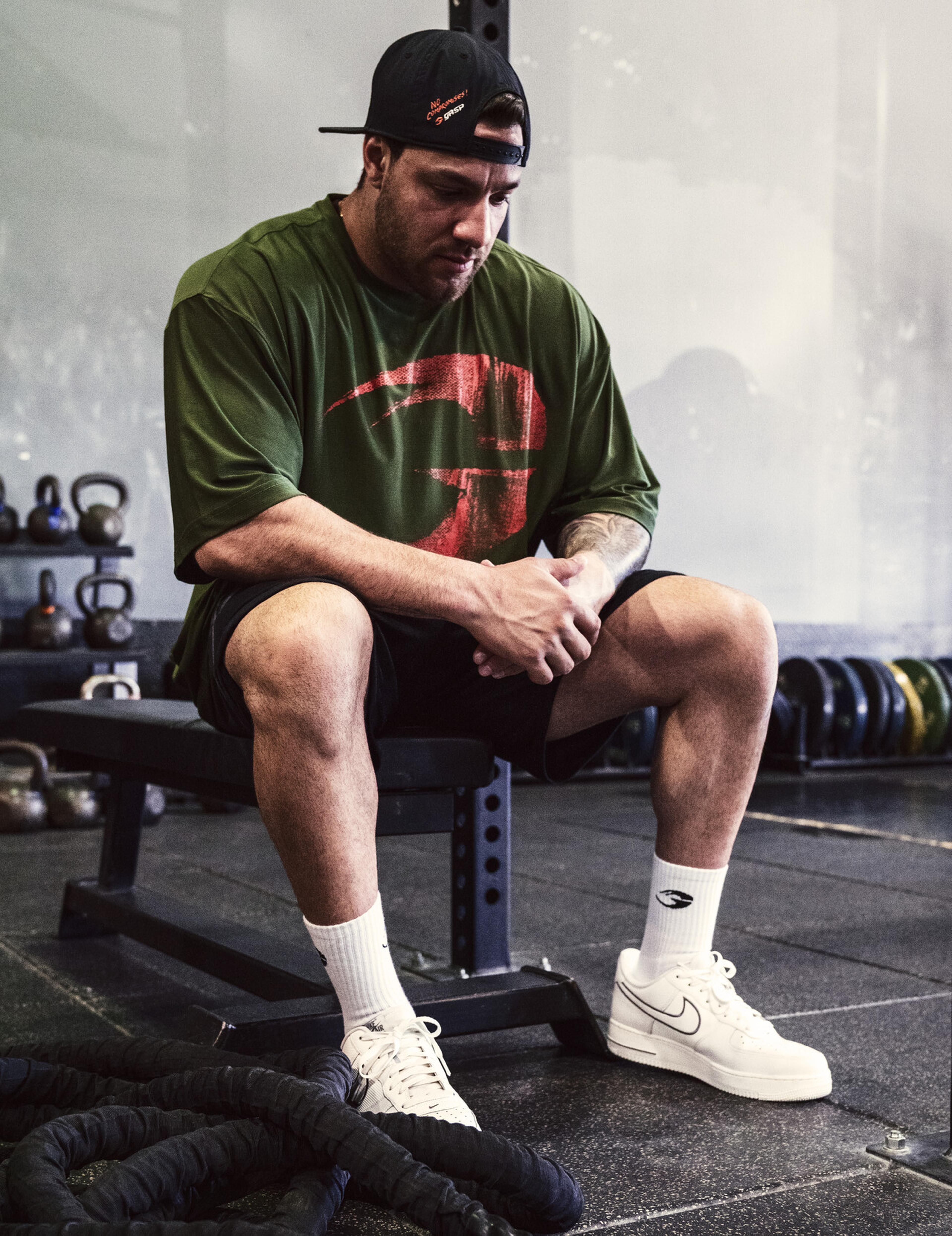 Man in a green shirt and cap sits on a gym bench, surrounded by kettlebells and ropes, looking pensive.
