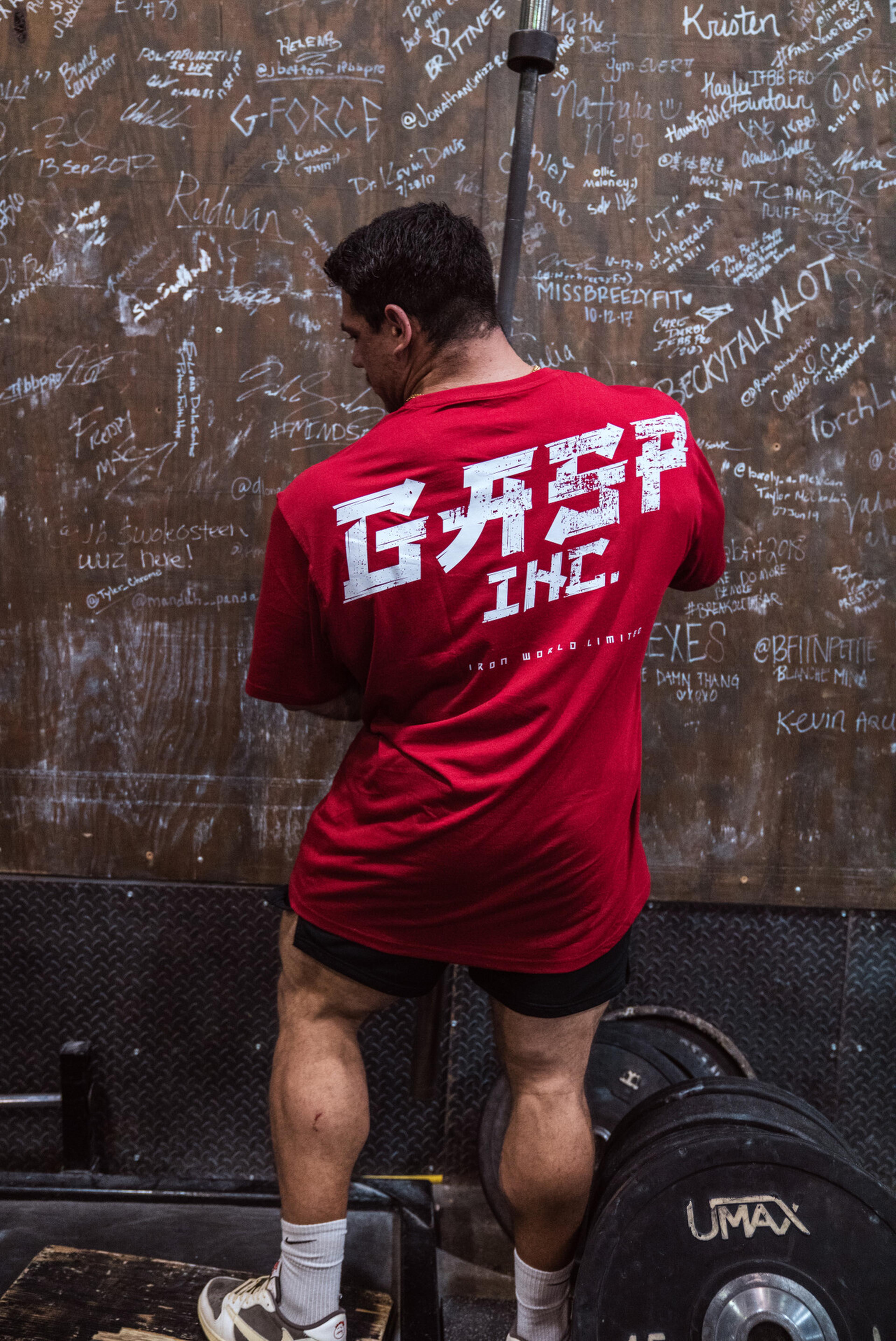 A person in a red "GRIP INC" shirt stands in front of a wall covered in signatures, next to a barbell and weights.