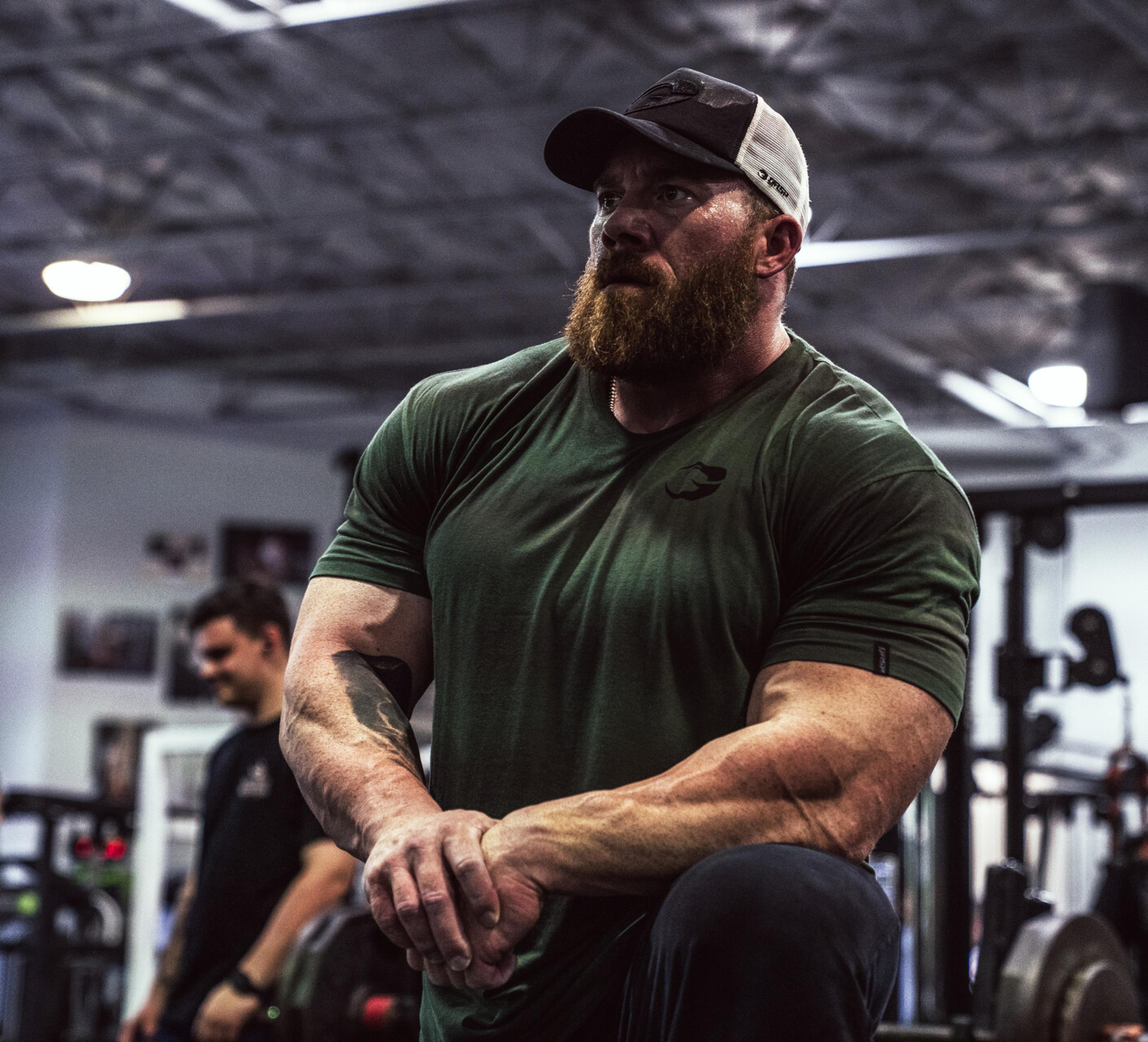 Bearded man in a green shirt and cap kneels in a gym, showing muscular arms. Gym equipment and another person are in the background.