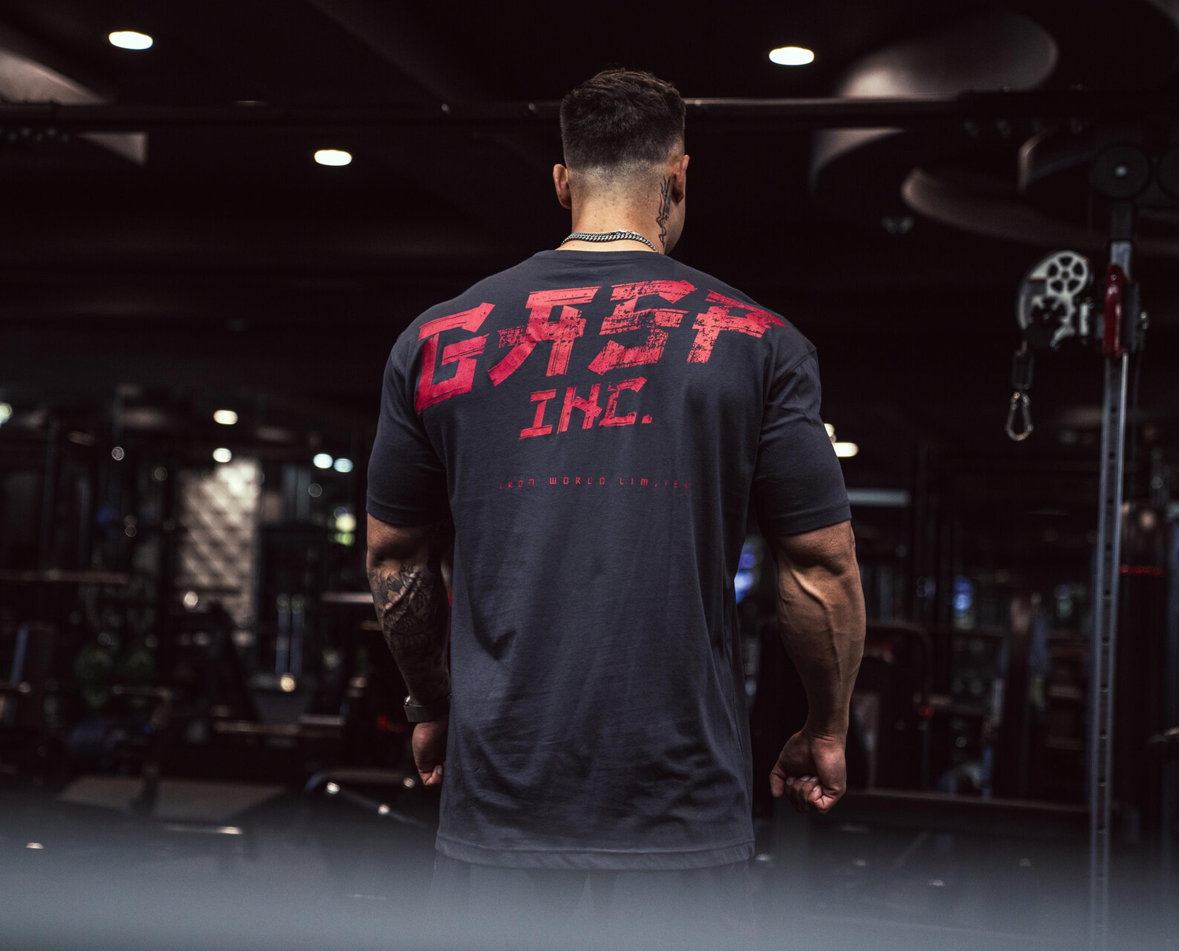 A muscular man in a gym wearing a black shirt with "GASP INC." in red letters on the back, standing near exercise equipment.